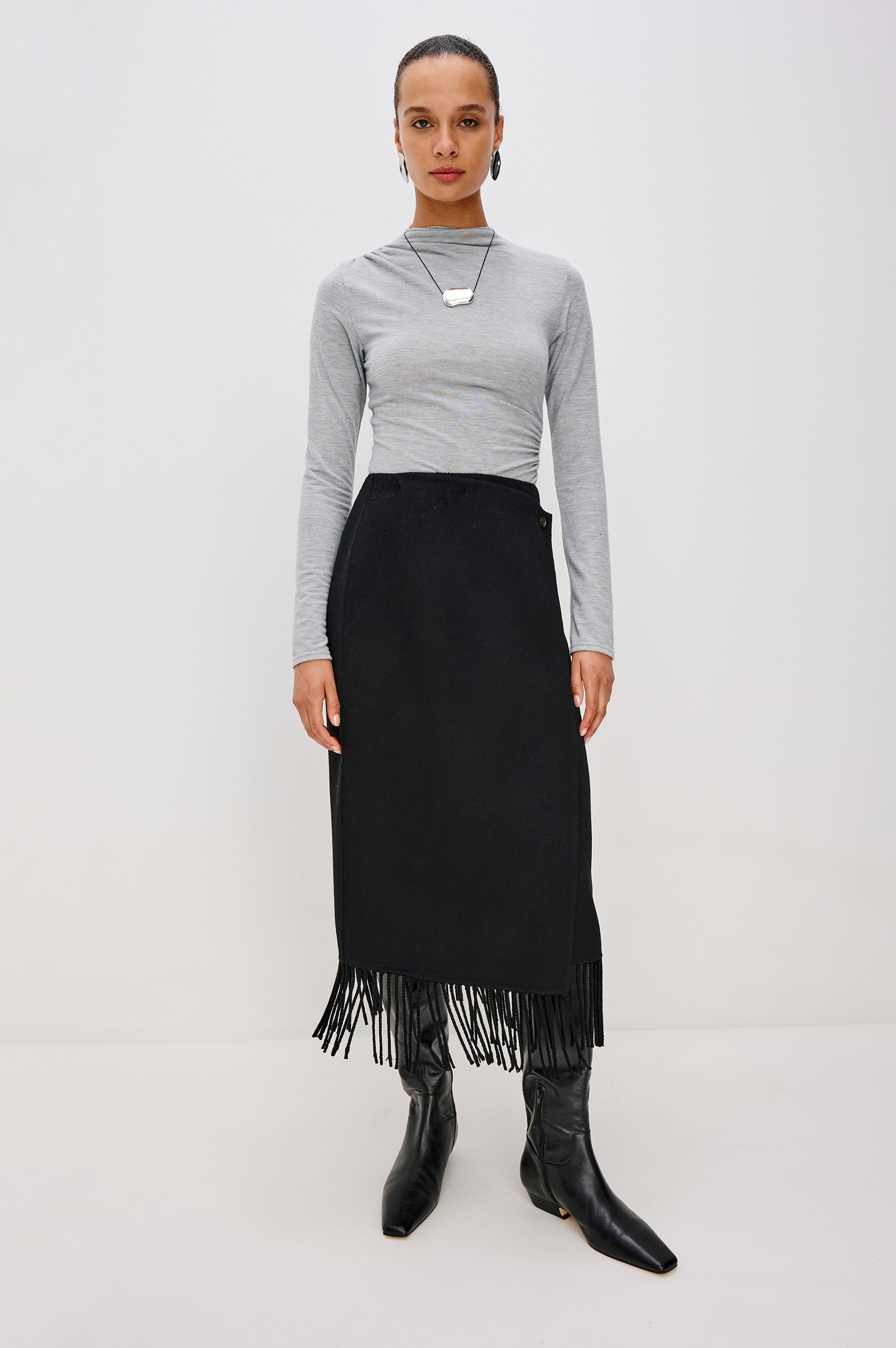 A high-waisted black wool-blend midi skirt. The wrap-style design fastens with a side button and interior closure, finished with statement fringe along the hem for added movement. Full Body Front View Featured on Model.