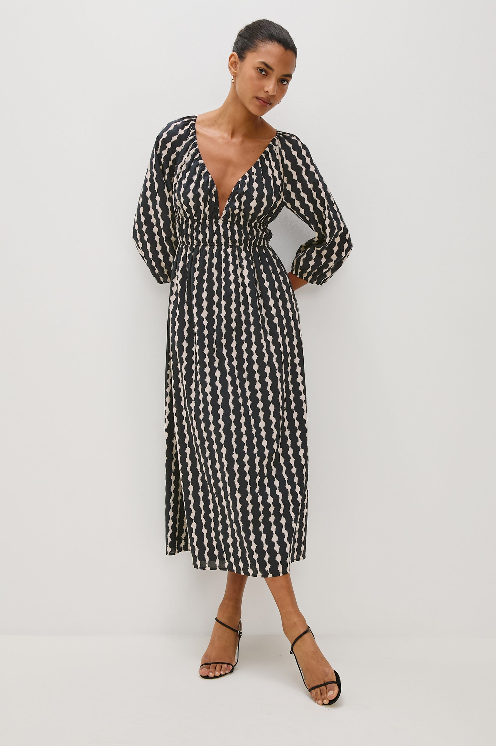 Paio midi dress with a fitted bodice, three quarter sleeves with a slight puff effect, flowing skirt, and eyelet closures at the chest in Painted Noir colorway - full body front view of model posing with hands behind back