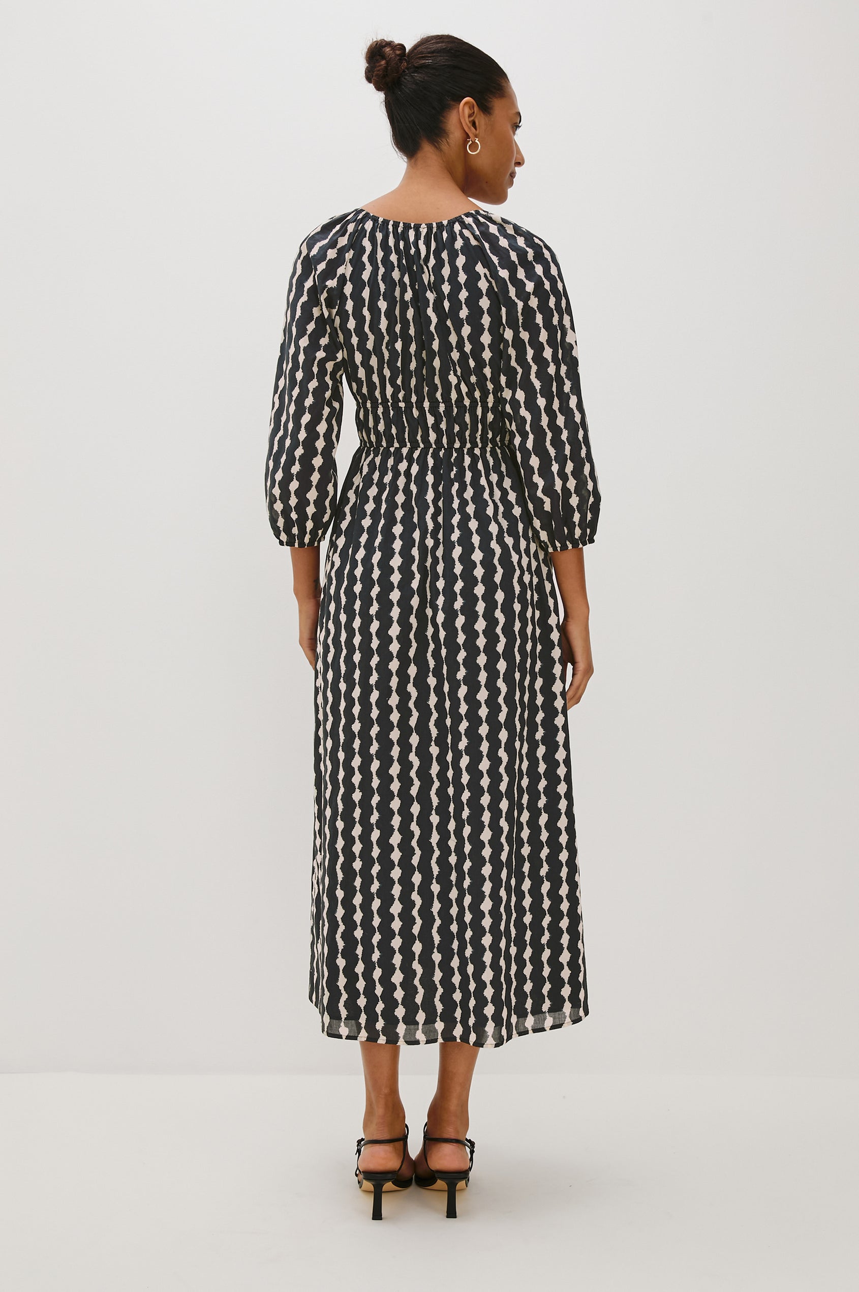 Paio midi dress with a fitted bodice, three quarter sleeves with a slight puff effect, flowing skirt, and eyelet closures at the chest in Painted Noir colorway - back full body view on model