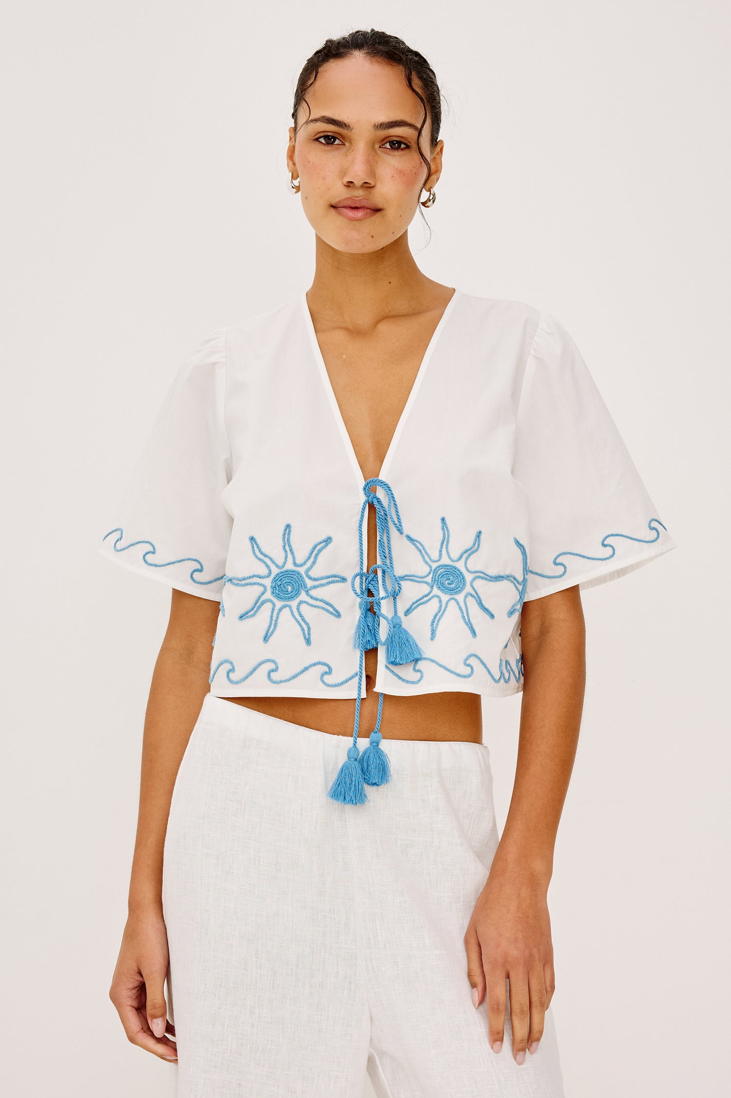 Pari Short-Sleeve Cropped Top with V Neck, Front Tie Closures, and Sun Embroidery Details in Blue Stitching Against White Fabric - Upper Body Front View Featured on Model