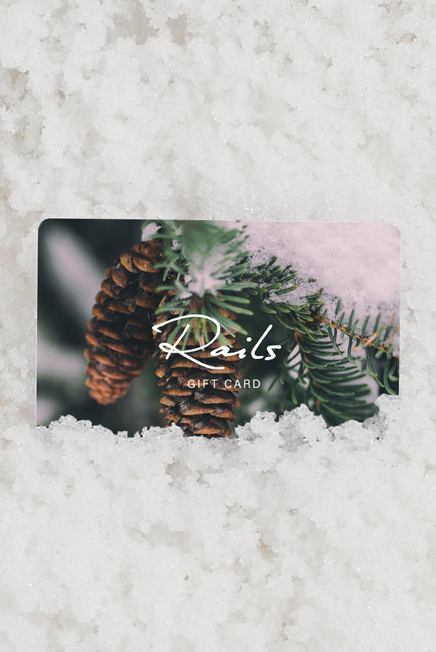Rails gift card partially tucked into fresh snow, featuring a close-up photo of pine cones hanging from a snow-dusted evergreen branch.