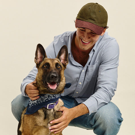 Smiling man crouching in a light blue button-up and olive cap, with his arms around a German Shepherd in a navy bandana.