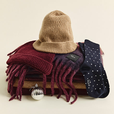Stack of folded scarves in burgundy, camel, and navy, topped with a camel ribbed beanie, a burgundy knit hat, and navy polka-dot socks, with a silver ornament at the base.