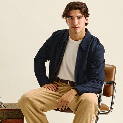 Seated man in a navy zip jacket over a cream tee, with tan trousers and a brown belt, on a tan leather chair.