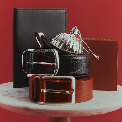 Black and cognac leather belts stacked with a black passport holder and a brown leather wallet on a marble table, with a silver ornament, against a red backdrop.