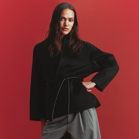 Woman in a black wool wrap jacket with a thin leather tie belt, wide-leg gray pleated trousers, and black strappy heeled sandals, against a red backdrop.