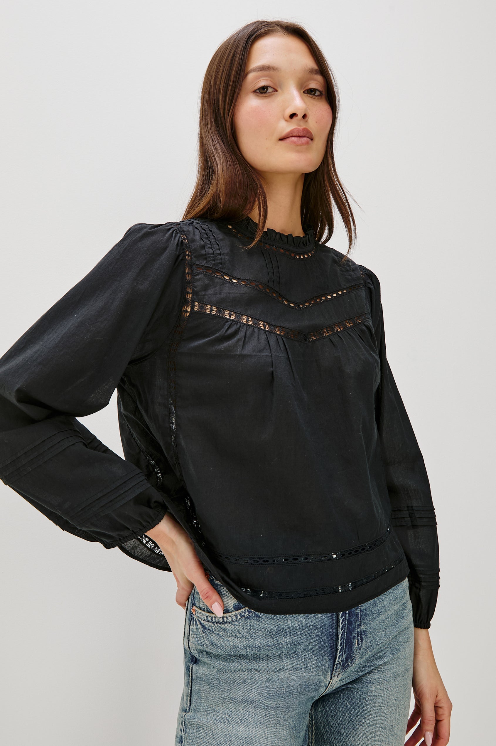 Front view of female model wearing our Reyna Top in black. This is a long sleeve crewneck baby doll blouse, with ruffles at the collar. Styled with light wash denim.