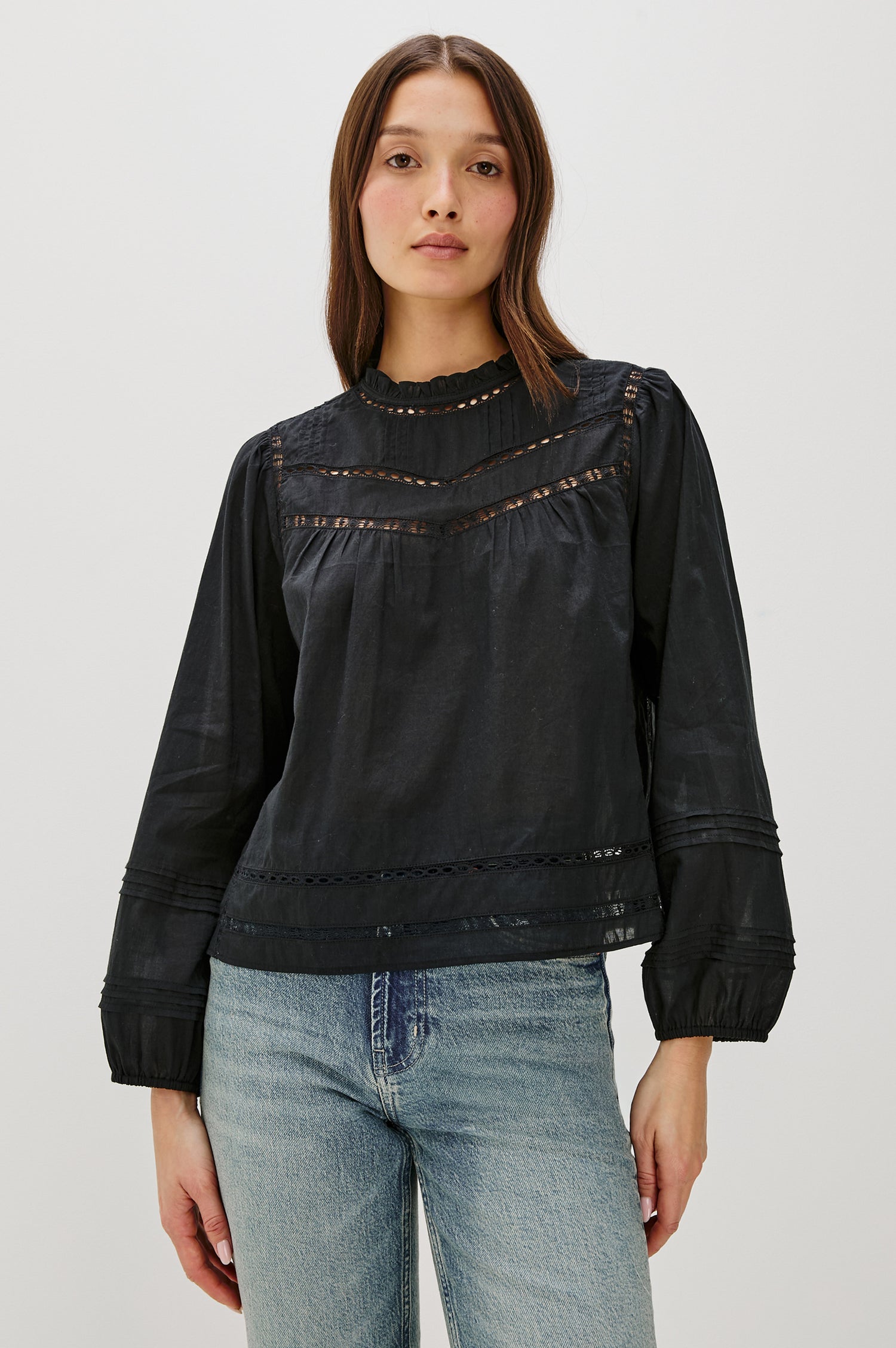 Front view of female model wearing our Reyna Top in black. This is a long sleeve crewneck baby doll blouse, with ruffles at the collar. Styled with light wash denim.