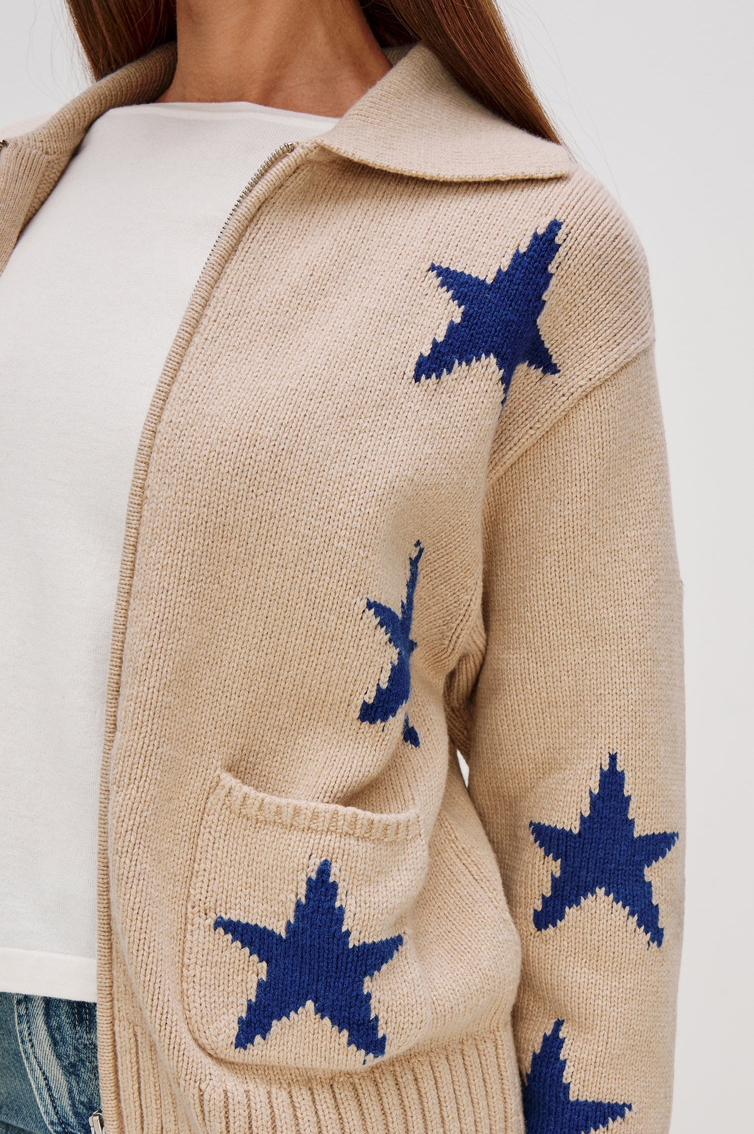 Rhea, a longline camel cardigan in a wool blend, featuring cobalt blue star motifs. The cardigan has a relaxed fit, zip-front closure, and front patch pockets. Close Up View Featured on Model, Unzipped