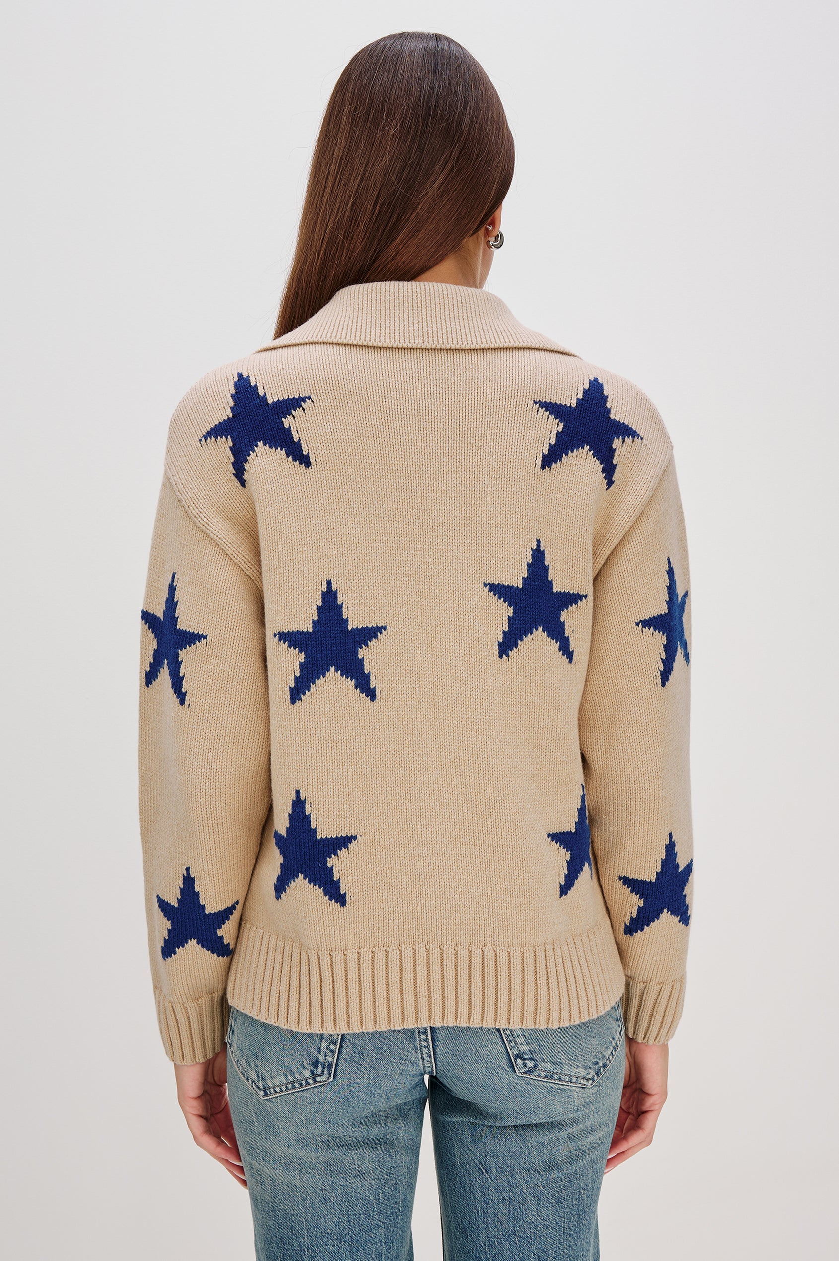 Rhea, a longline camel cardigan in a wool blend, featuring cobalt blue star motifs. The cardigan has a relaxed fit, zip-front closure, and front patch pockets. Back View Featured on Model, Zipped Up