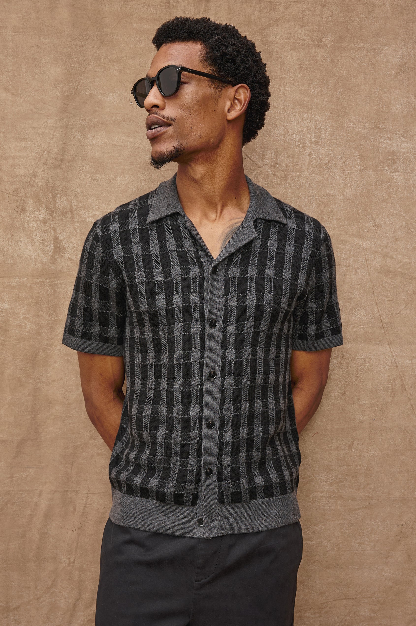 The Ronan is a collared button front knit shirt with short sleeves, grey rib trim, grey + black block stitch pattern in Charcoal Black colorway. Front view with sunglasses featured on model.