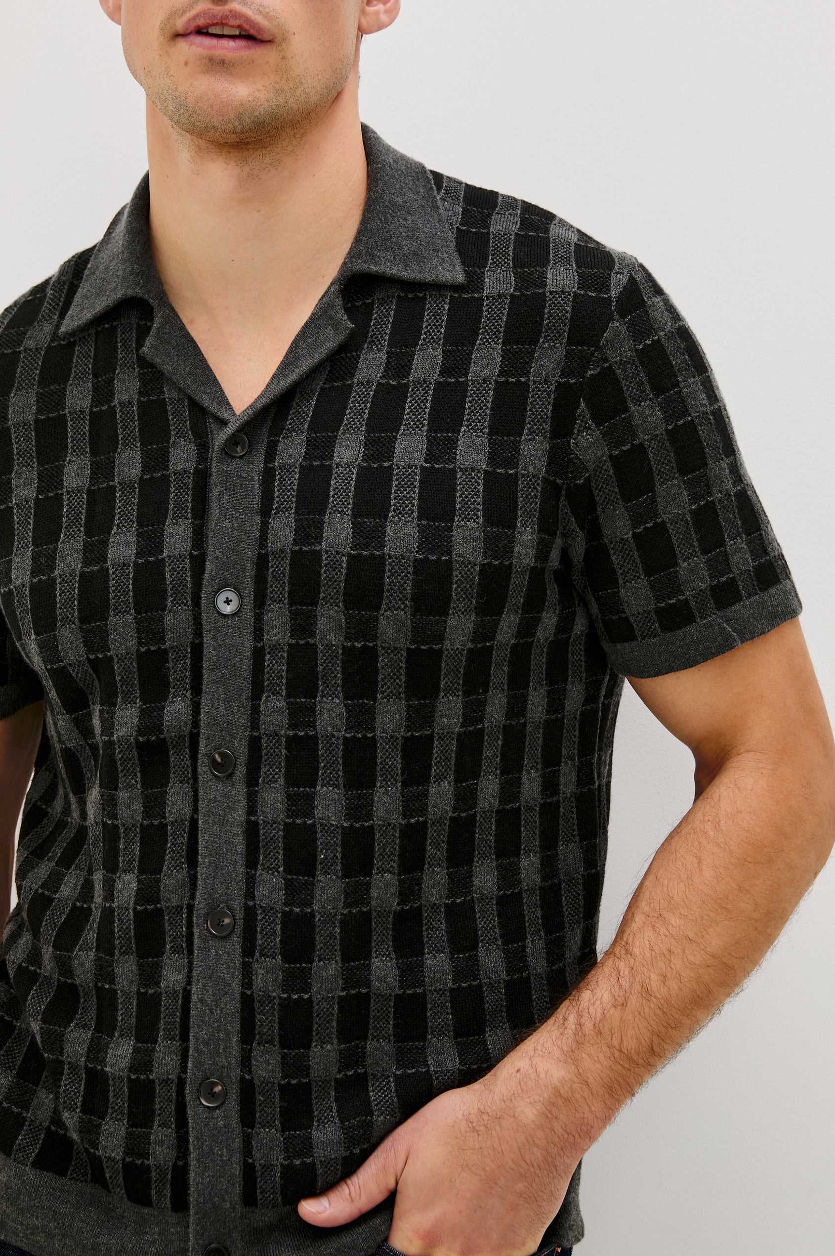 The Ronan is a collared button front knit shirt with short sleeves, grey rib trim, grey + black block stitch pattern in Charcoal Black colorway. Front detail view featured on model.