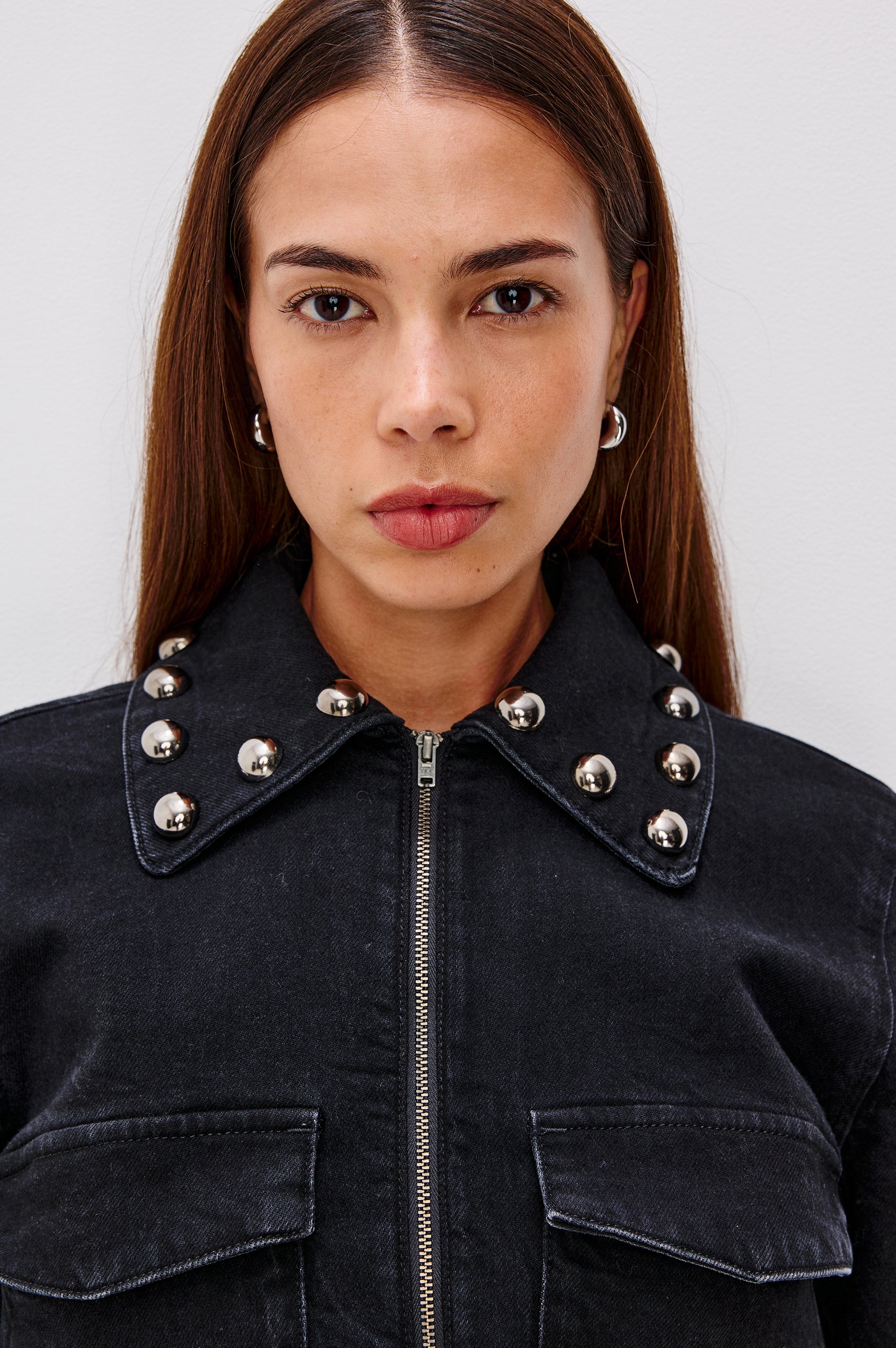 Roxbury is a boxy fit denim jacket featuring nail-back studs at the oversized collar, cuffs, and hem. It has oversized chest pockets with hidden snap closures, hem tabs, and a sleek front zipper closure in Black Stone Studded colorway. Front detail view featured on model.