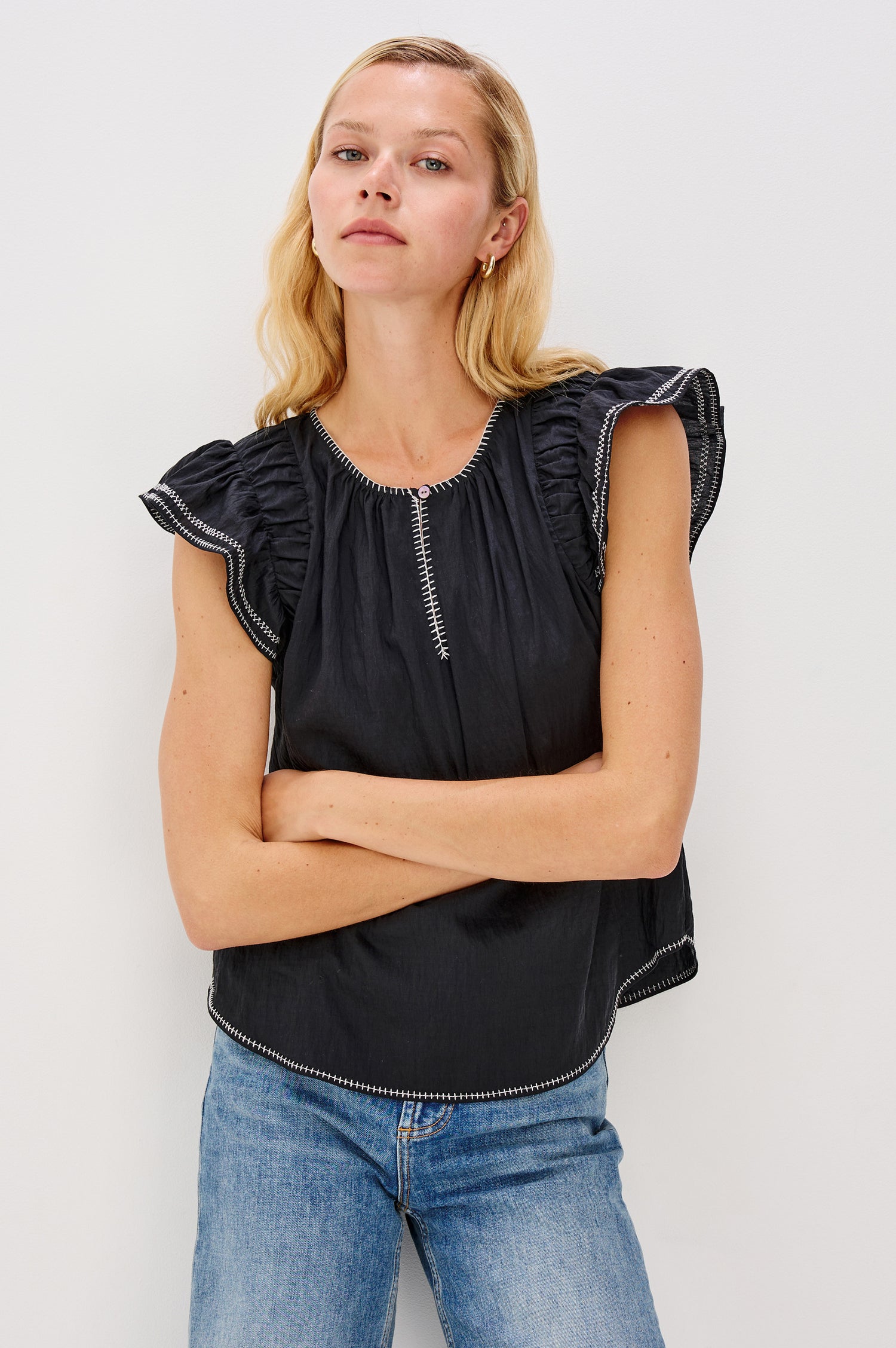 Sable is a scoop neck top with flutter sleeves and a split neck that is adjustable with a button, it has a high low hem and contrast white stitching in Black colorway. Front view featured on model.