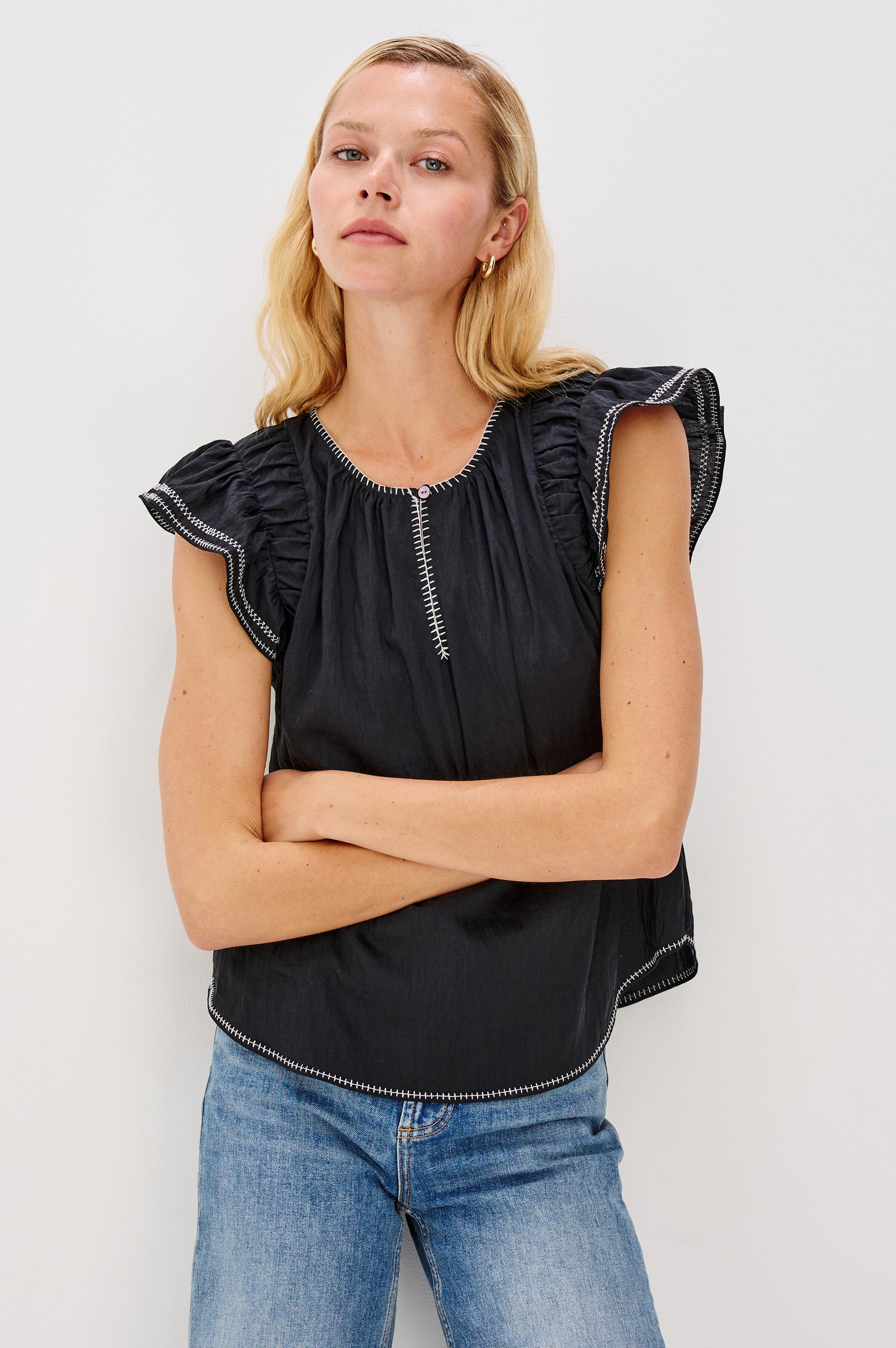 Sable is a scoop neck top with flutter sleeves and a split neck that is adjustable with a button, it has a high low hem and contrast white stitching in Black colorway. Front view featured on model.