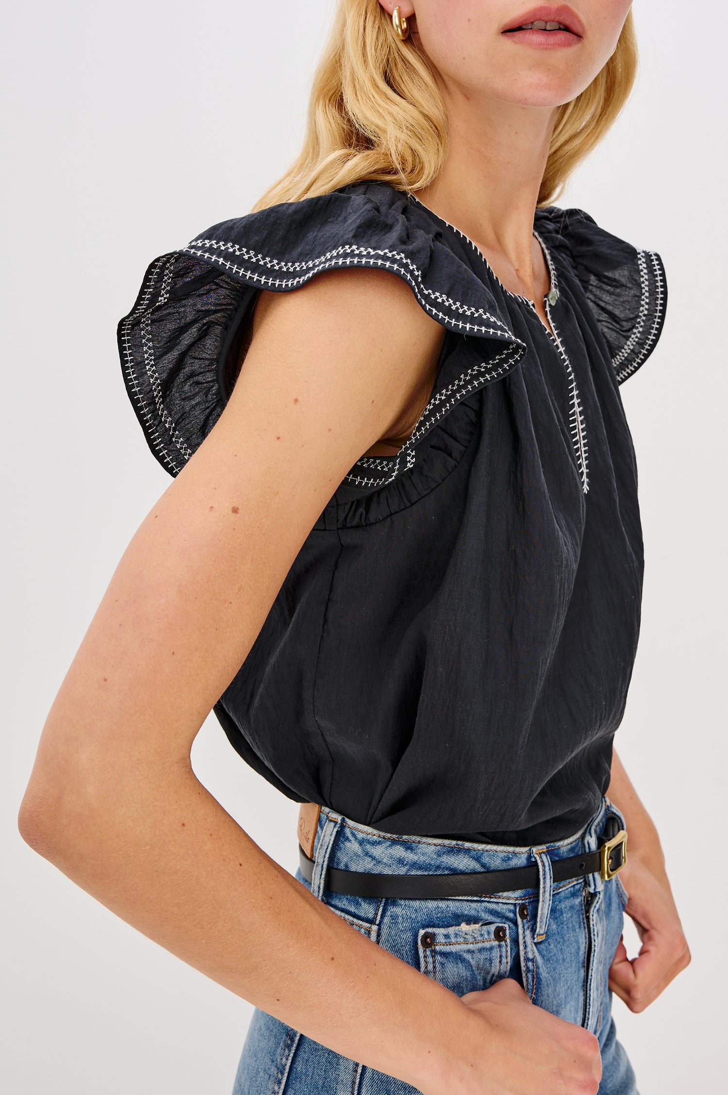 Sable is a scoop neck top with flutter sleeves and a split neck that is adjustable with a button, it has a high low hem and contrast white stitching in Black colorway. Side detail view featured on model.
