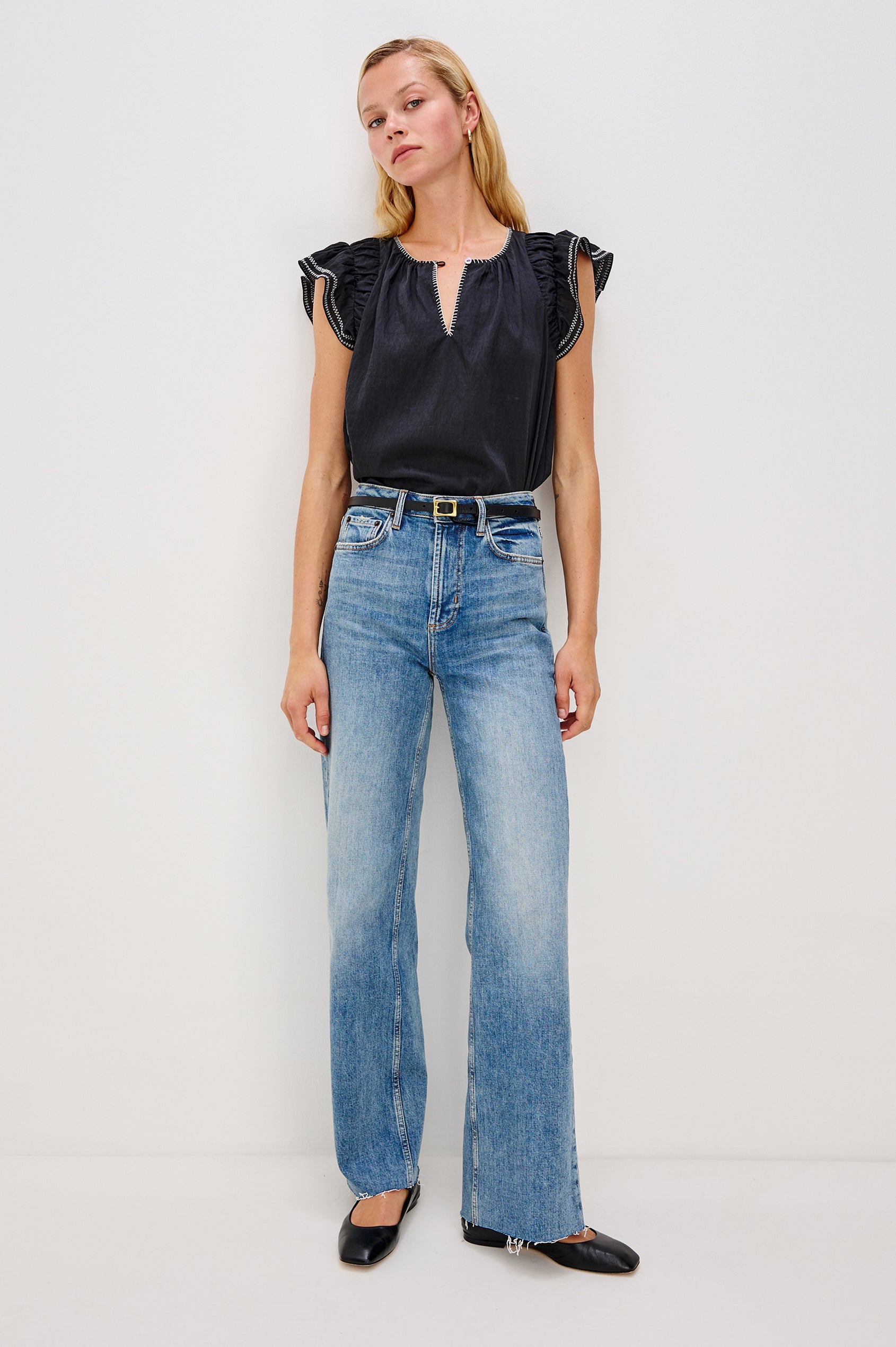 Sable is a scoop neck top with flutter sleeves and a split neck that is adjustable with a button, it has a high low hem and contrast white stitching in Black colorway. Front full body view featured on model.