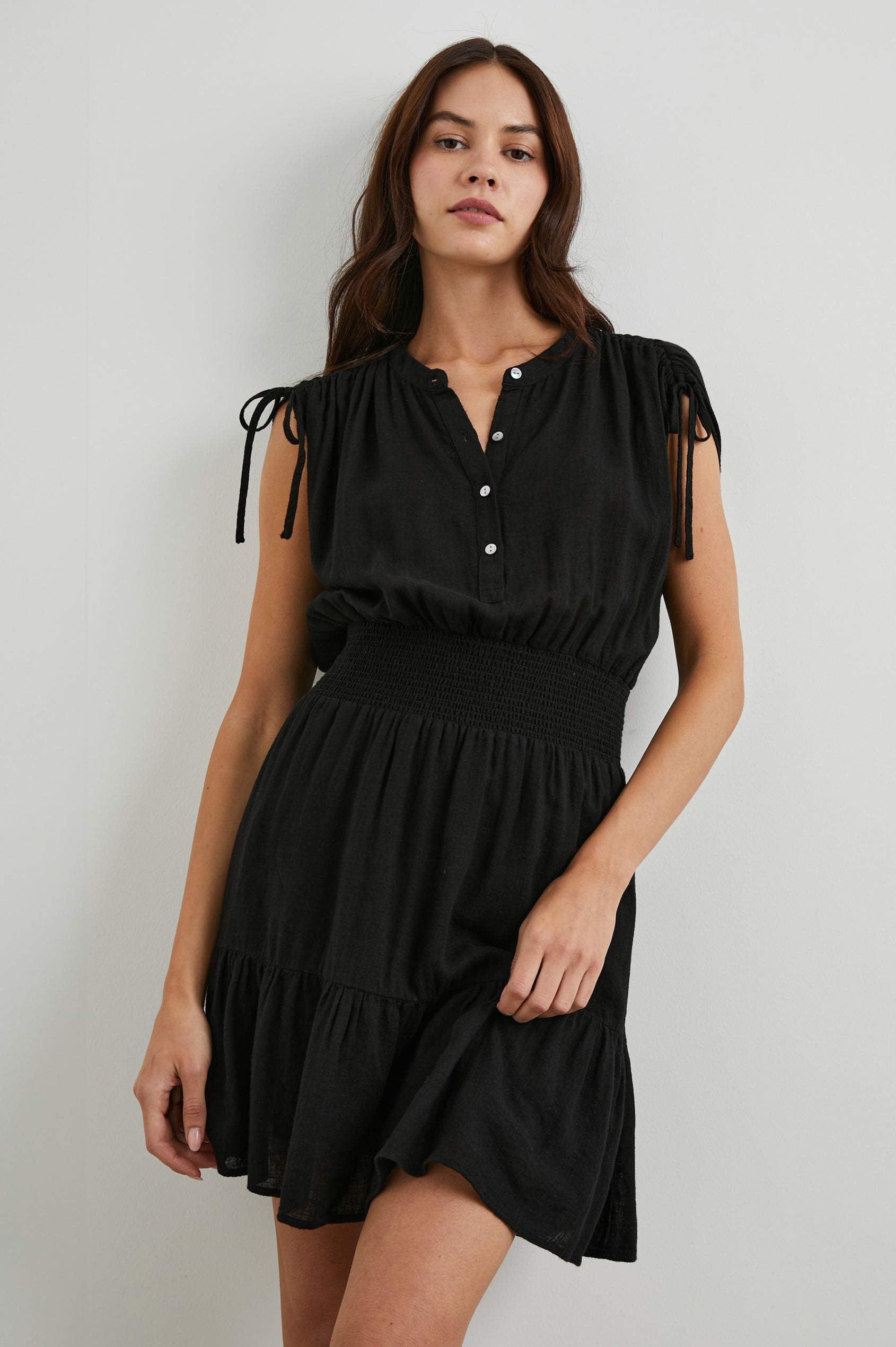 Samina Above-Knee Sleeveless Dress with drawstrings at the Sleeve Openings, Buttons along the Bodice, A Cinched Waist and a Tiered Skirt in Black Colorway - Front View Featured on Model 