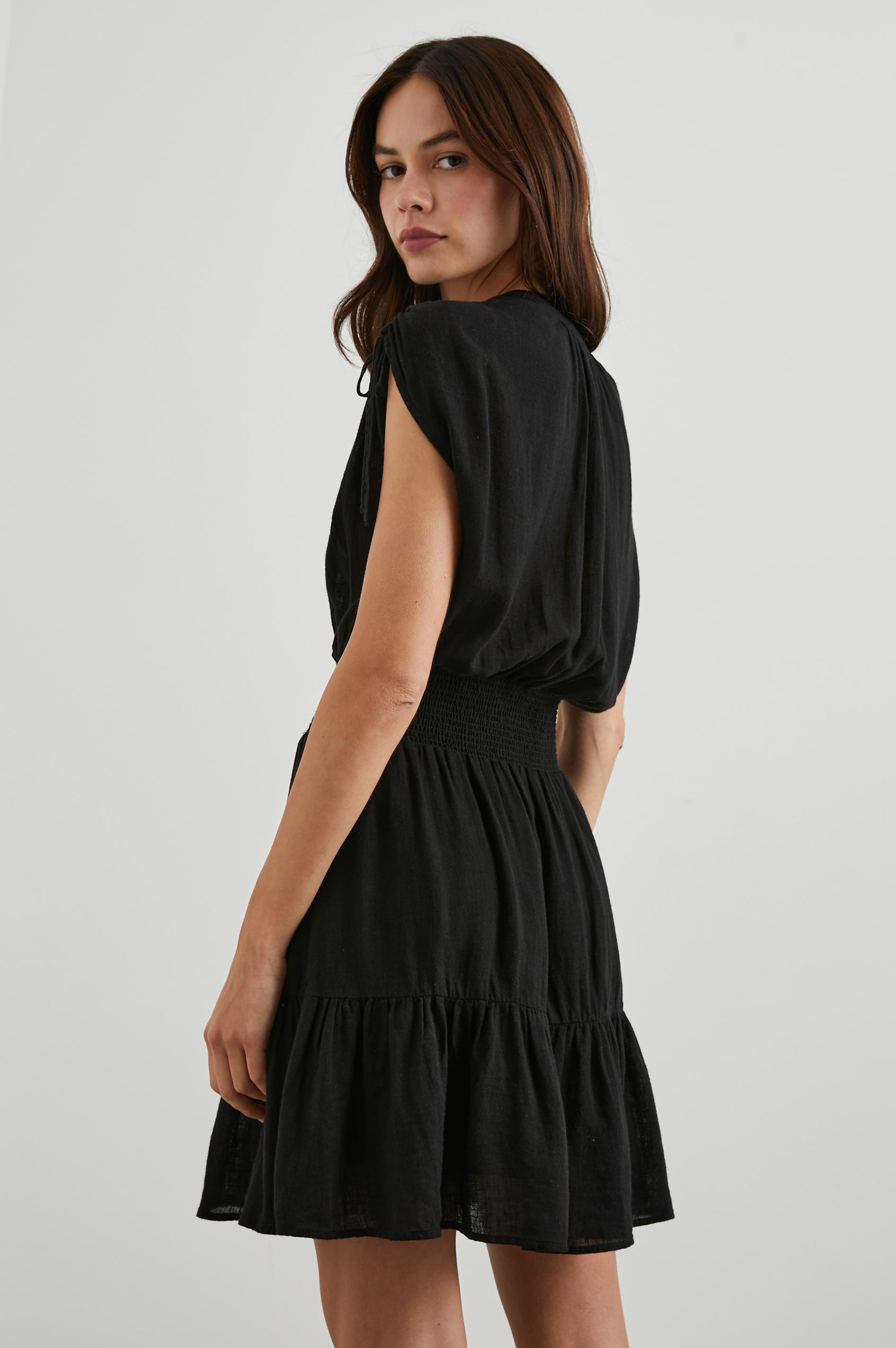 Samina Above-Knee Sleeveless Dress with drawstrings at the Sleeve Openings, Buttons along the Bodice, A Cinched Waist and a Tiered Skirt in Black Colorway - Back View Featured on Model 