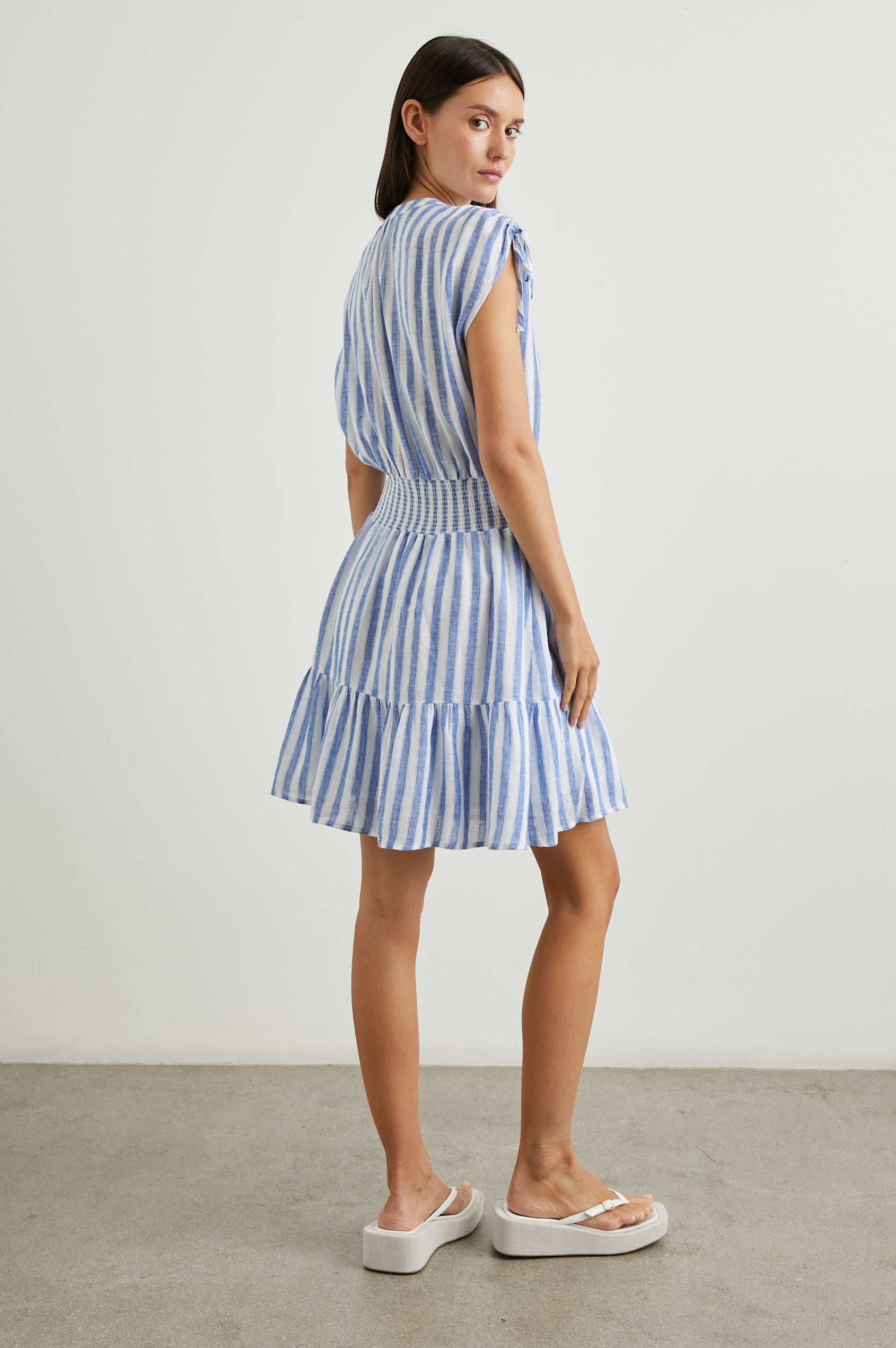 Samina Above-Knee Sleeveless Dress with drawstrings at the Sleeve Openings, Buttons along the Bodice, A Cinched Waist and a Tiered Skirt in Casablanca Stripe Colorway, with blue vertical stripes on white - Back Full Body View Featured on Model 