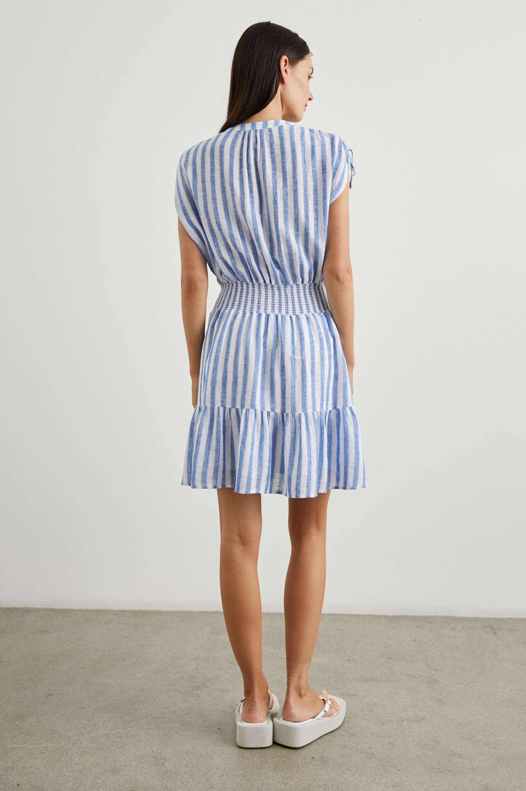 Samina Above-Knee Sleeveless Dress with drawstrings at the Sleeve Openings, Buttons along the Bodice, A Cinched Waist and a Tiered Skirt in Casablanca Stripe Colorway, with blue vertical stripes on white - Back Full Body View Featured on Model 