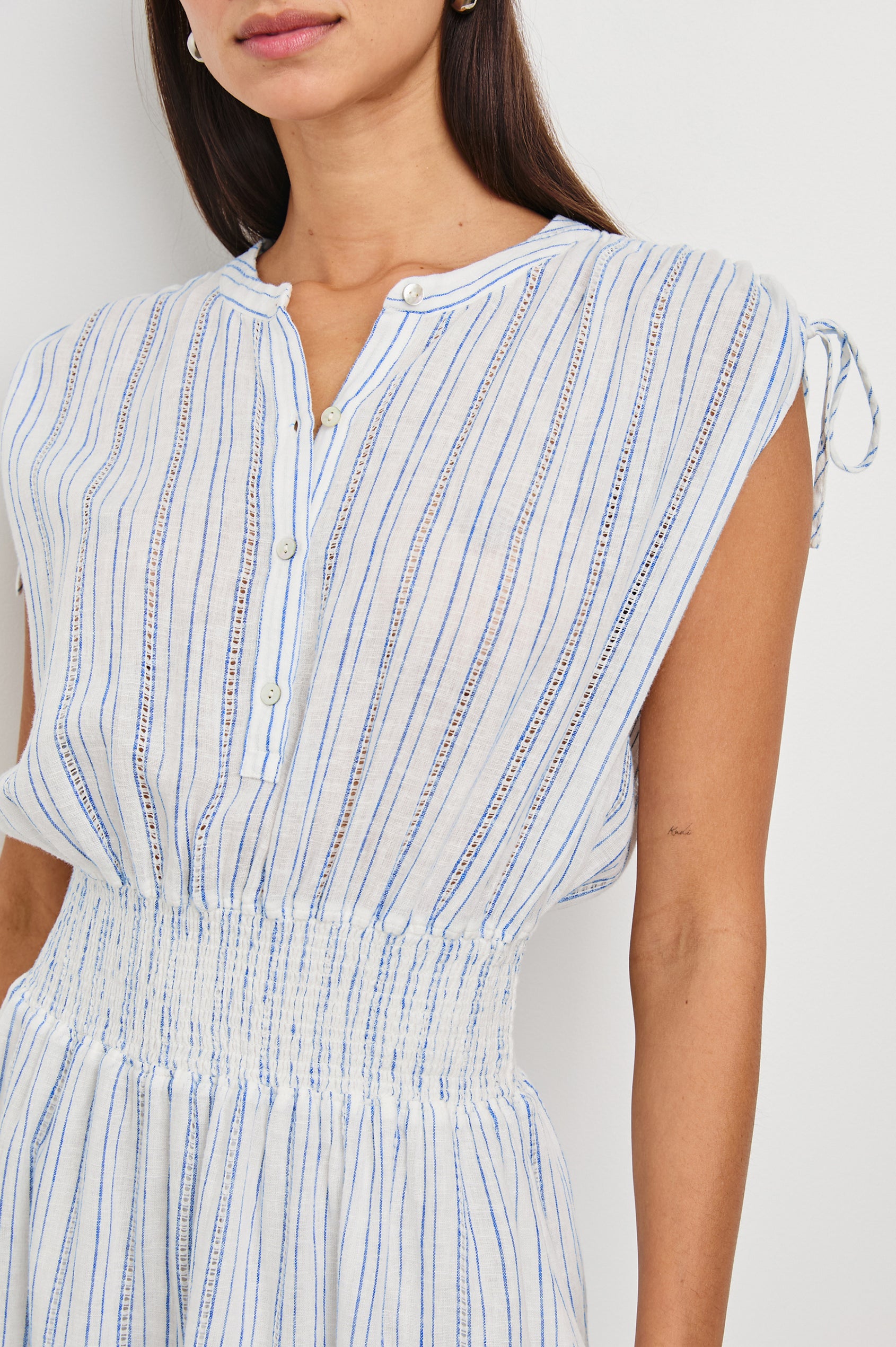 Samina Above-Knee Sleeveless Dress with drawstrings at the Sleeve Openings, Buttons along the Bodice, A Cinched Waist and a Tiered Skirt in Sonoma Stripe Colorway, Thin Vertical Blue Stripes on White - Front View Featured on Model 