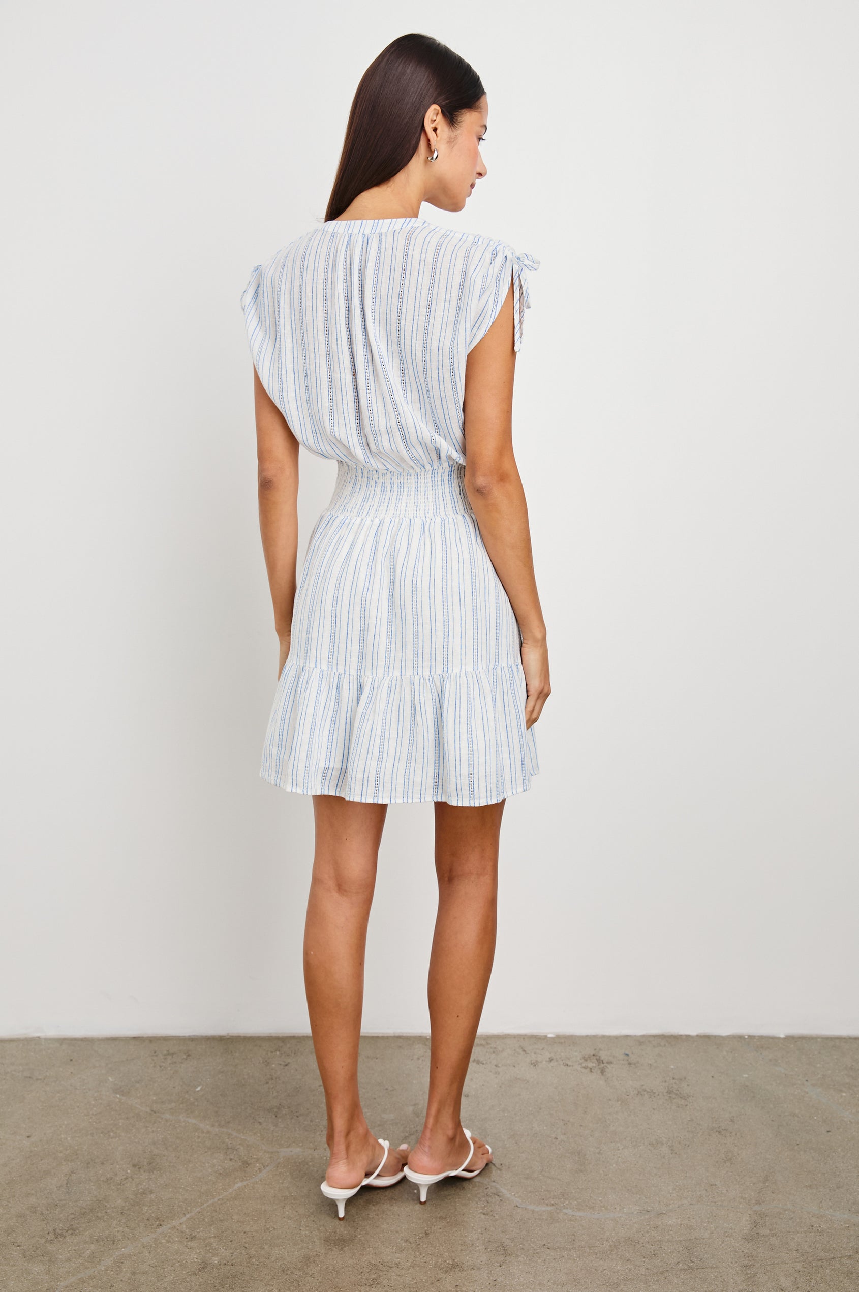 Samina Above-Knee Sleeveless Dress with drawstrings at the Sleeve Openings, Buttons along the Bodice, A Cinched Waist and a Tiered Skirt in Sonoma Stripe Colorway, Thin Vertical Blue Stripes on White - Back Full Body View Featured on Model 