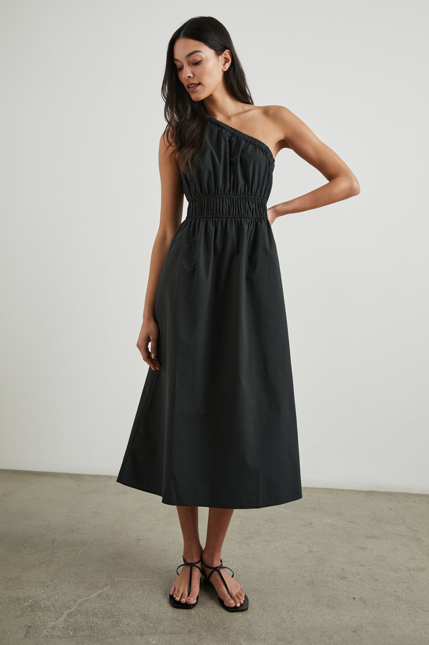 Selani Midi Dress With an Asymmetrical Neckline, Mid-Thigh Slide Slit and Cinched Waistband in Black Colorway - Front Full Body View Featured on Model