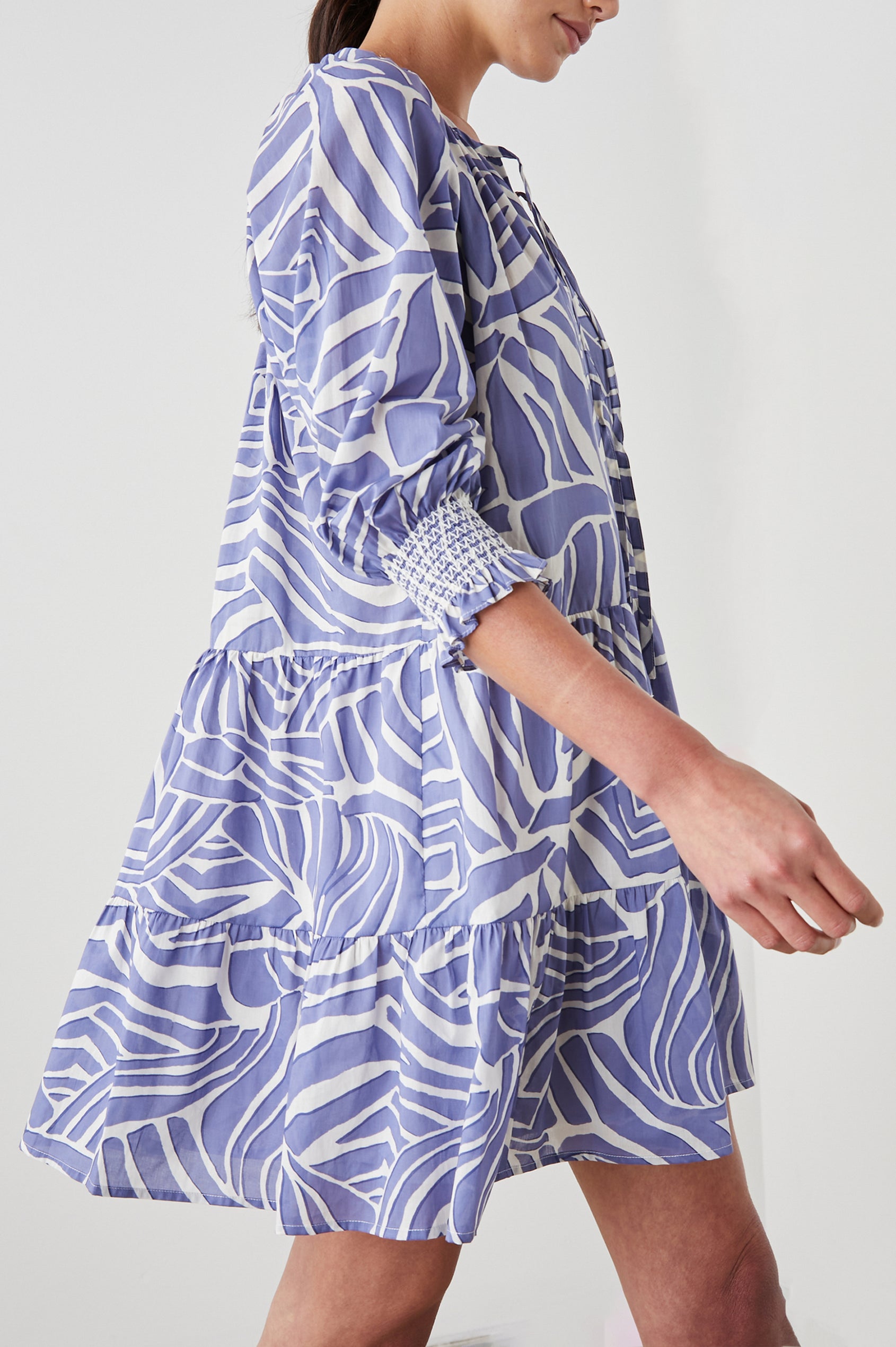 Sia Mini Dress with Three-Quarter Sleeves, Shirred Cuffs, a Tiered Skirt, and a V Neck with a Tie and Light Ruching in Island Waves Colorway, a Purple and White Print - Side View Featured on Model 