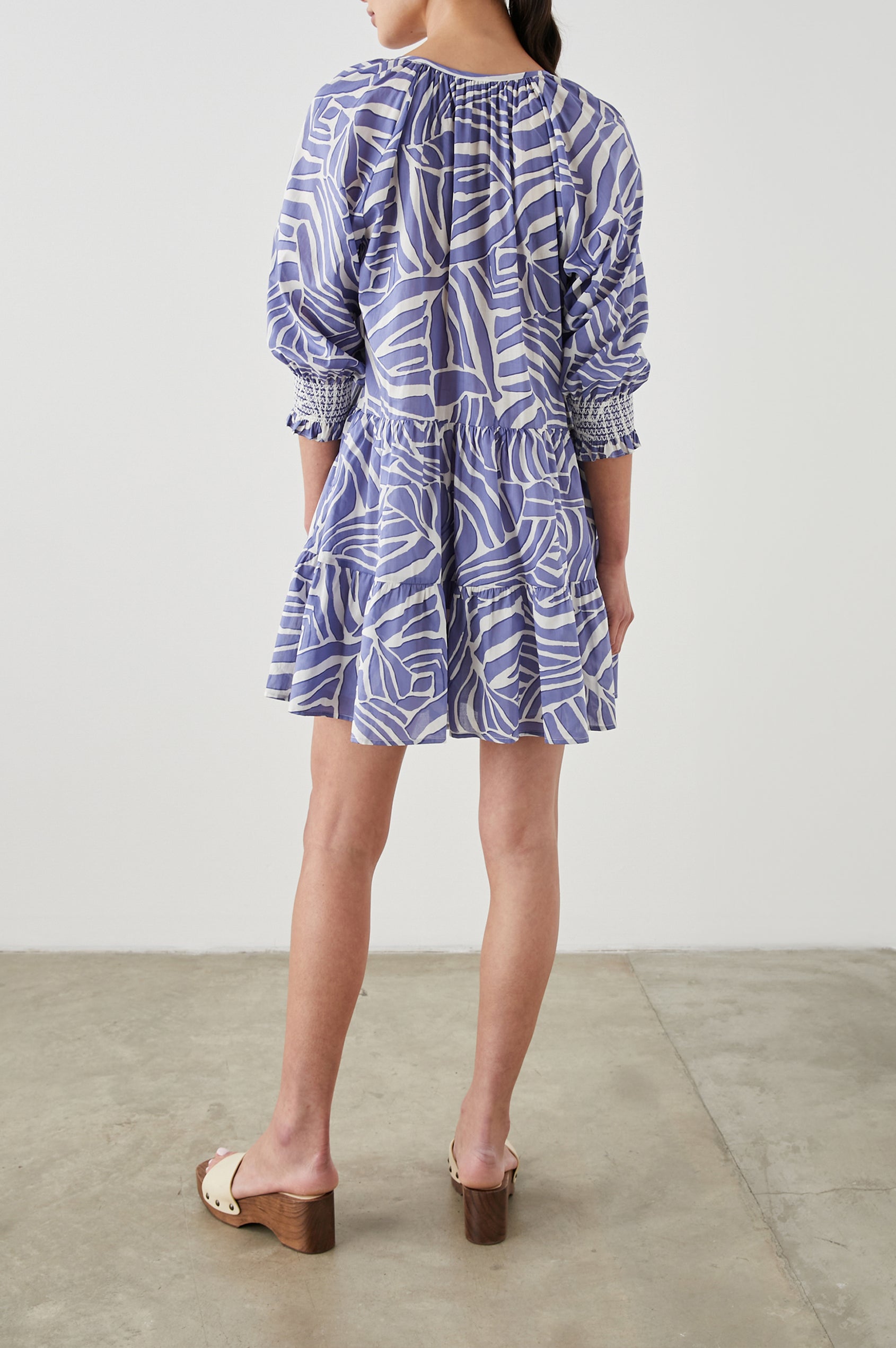 Sia Mini Dress with Three-Quarter Sleeves, Shirred Cuffs, a Tiered Skirt, and a V Neck with a Tie and Light Ruching in Island Waves Colorway, a Purple and White Print - Back Full Body View Featured on Model 
