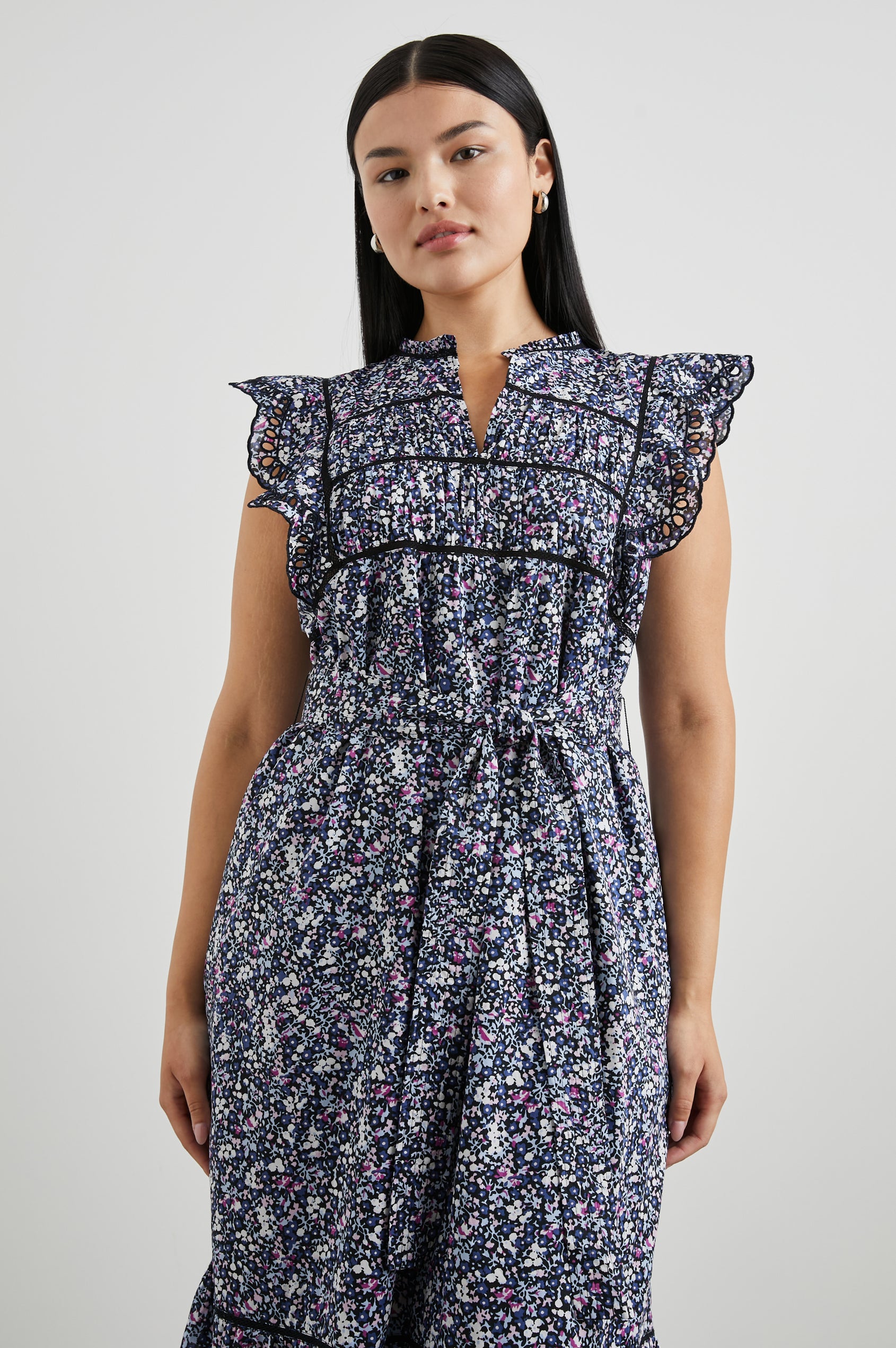 Sofie Short Sleeve Midi Dress with Flutter Sleeves, a V Neck, Panels with Shirring on the Bodice, a Fabric Waist Tie, a Tiered Skirt, and Cutouts on the Sleeves and Along the Hem in Midnight Hyacinth Colorway, Small Flowers in Blues, Pinks and White on Midnight - Front View Featured on Model 