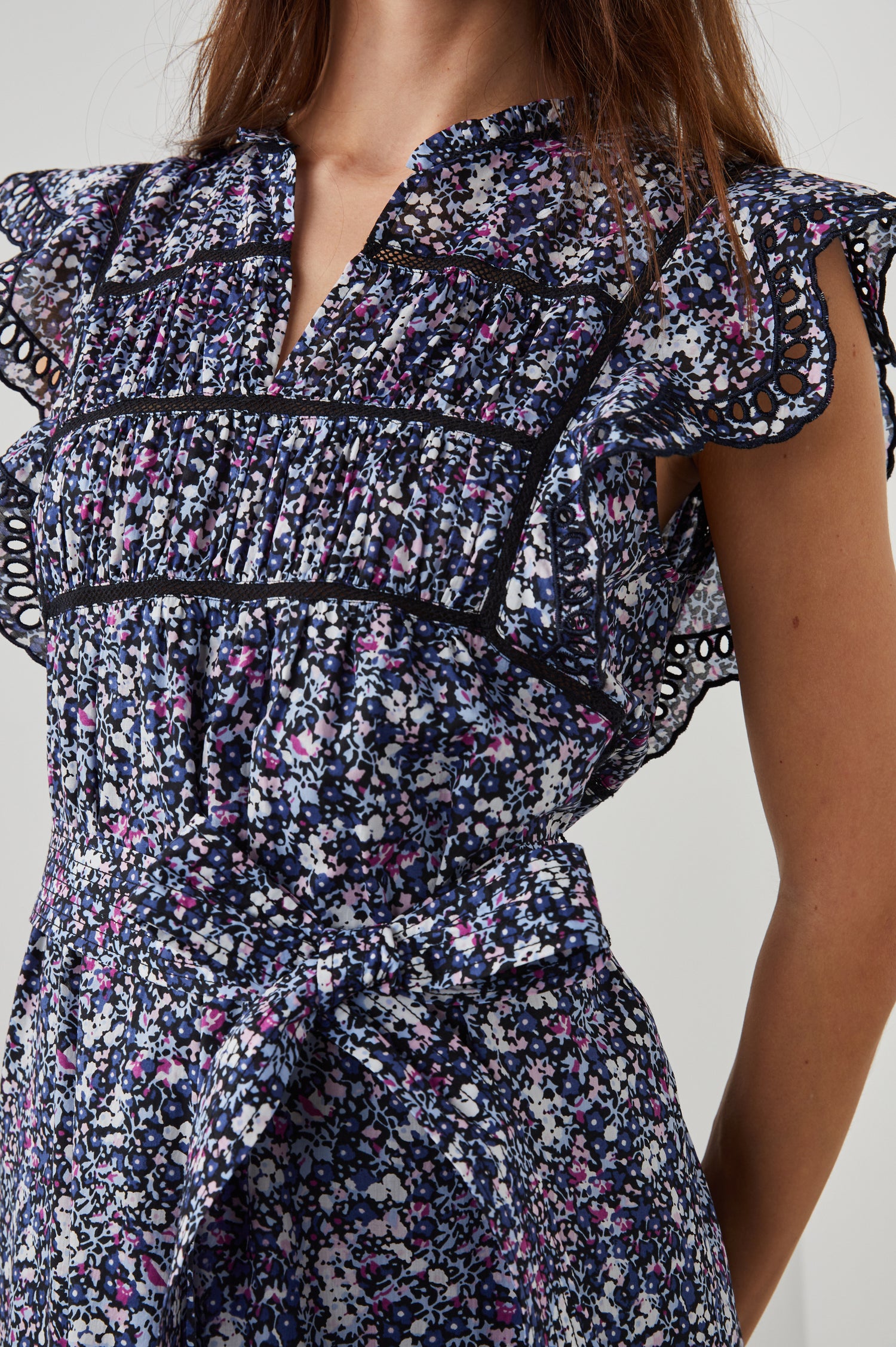 Sofie Short Sleeve Midi Dress with Flutter Sleeves, a V Neck, Panels with Shirring on the Bodice, a Fabric Waist Tie, a Tiered Skirt, and Cutouts on the Sleeves and Along the Hem in Midnight Hyacinth Colorway, Small Flowers in Blues, Pinks and White on Midnight - Front View Featured on Model 