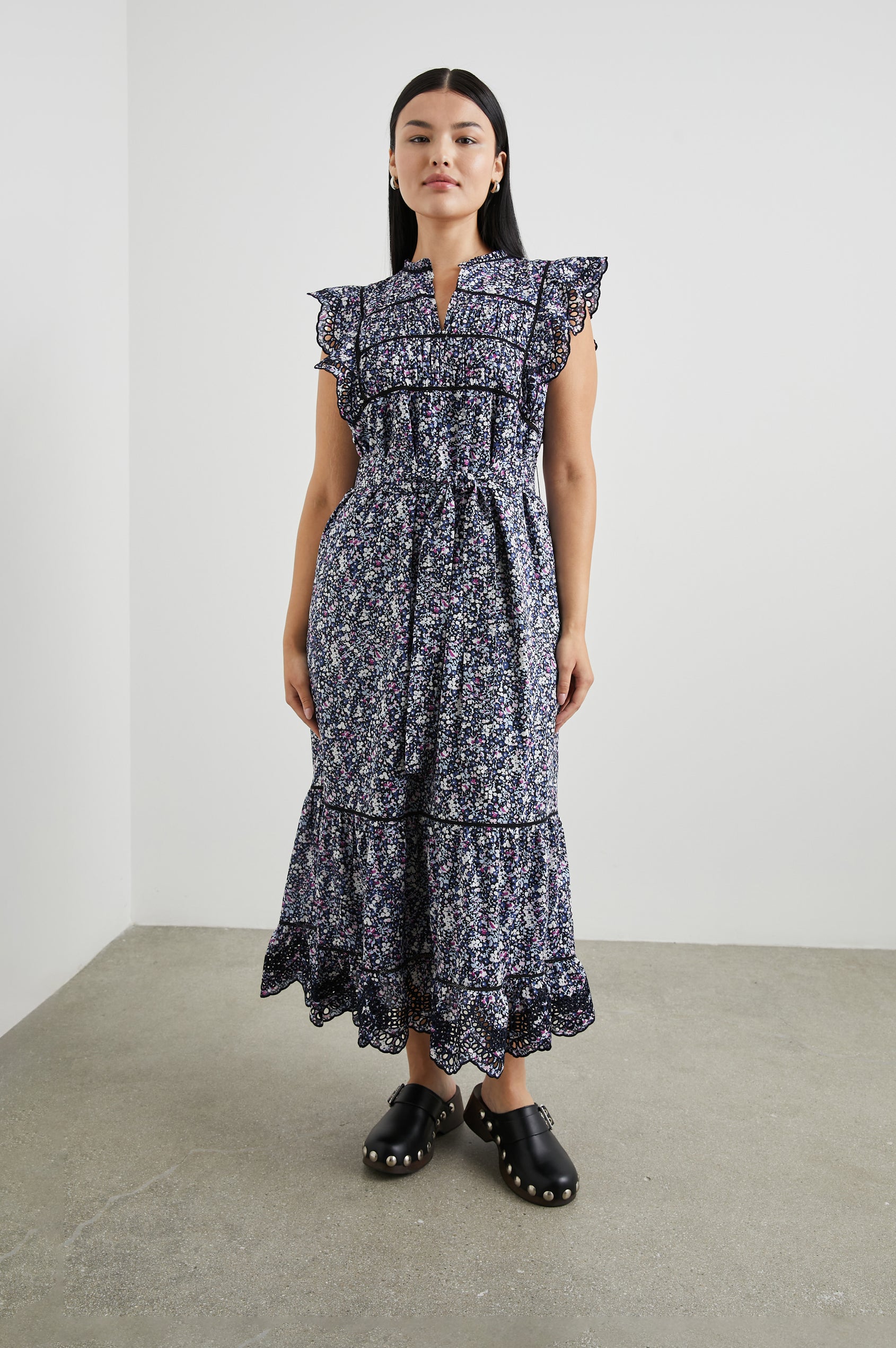 Sofie Short Sleeve Midi Dress with Flutter Sleeves, a V Neck, Panels with Shirring on the Bodice, a Fabric Waist Tie, a Tiered Skirt, and Cutouts on the Sleeves and Along the Hem in Midnight Hyacinth Colorway, Small Flowers in Blues, Pinks and White on Midnight - Front Full Body View Featured on Model 