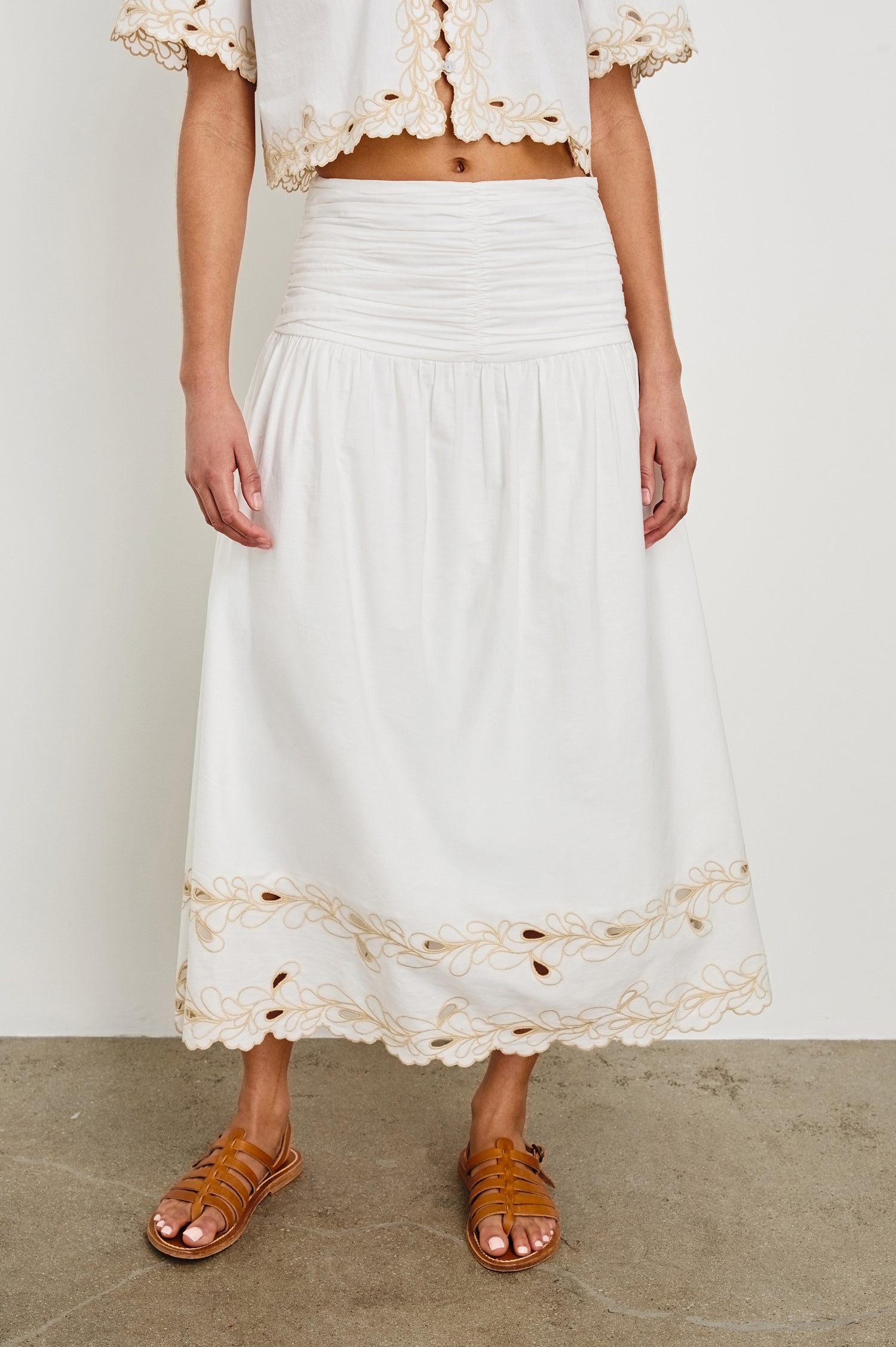 Detail image of model wearing a white, cotton poplin midi skirt with a lightly gathered waist that cinches and drops at the hips. Details include beige embroidered eyelets at the hem. Worn with brown sandals.