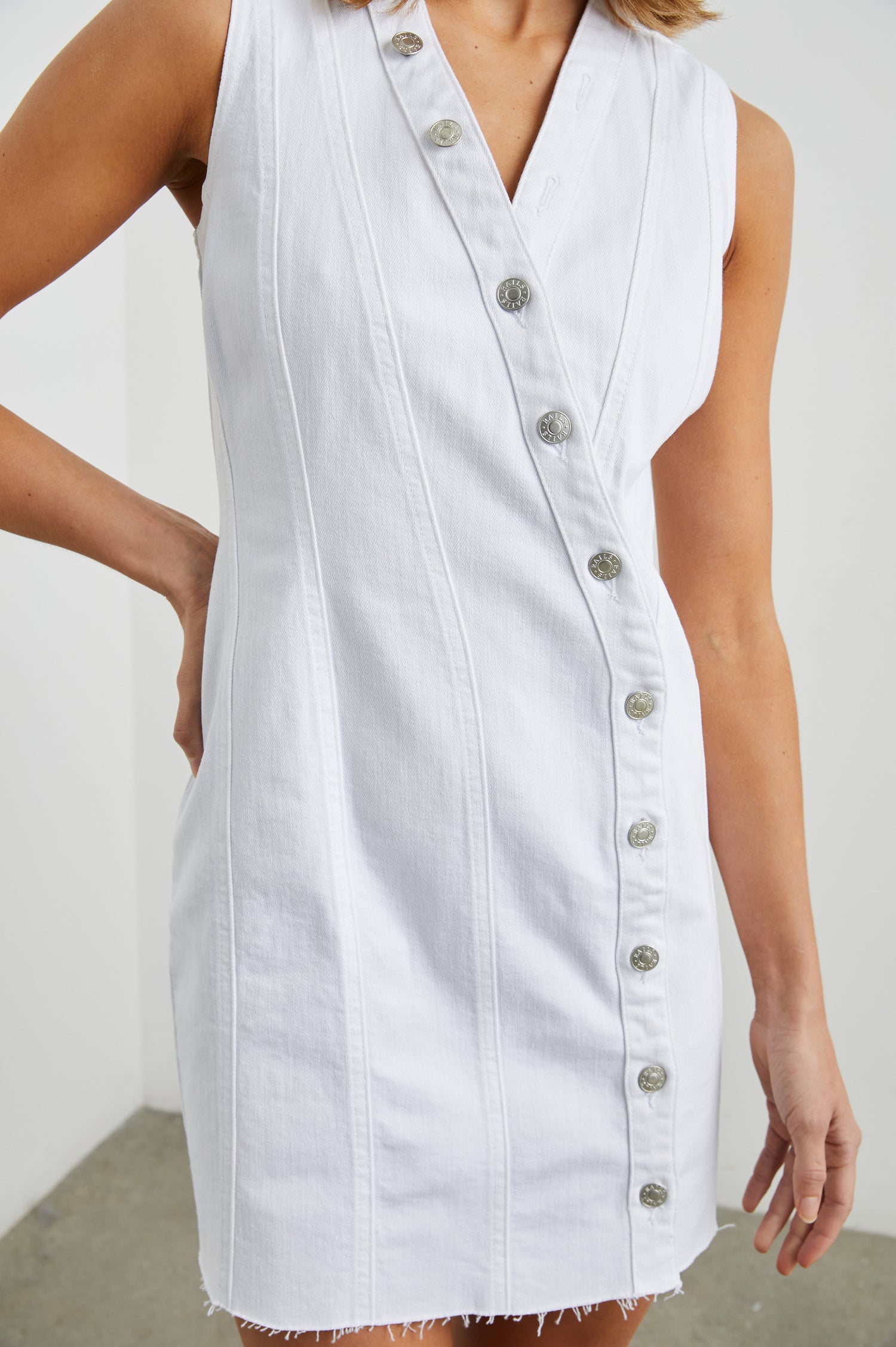 Strand Sleeveless Denim Mini Dress with a V Neck, a Raw Hem, Wide Shoulder Straps, a Row of Buttons that Extend Diagonally Across the Bodice and then Vertically Down the Skirt, Vertical Seams in Salt White Colorway - Front View Featured on Model  