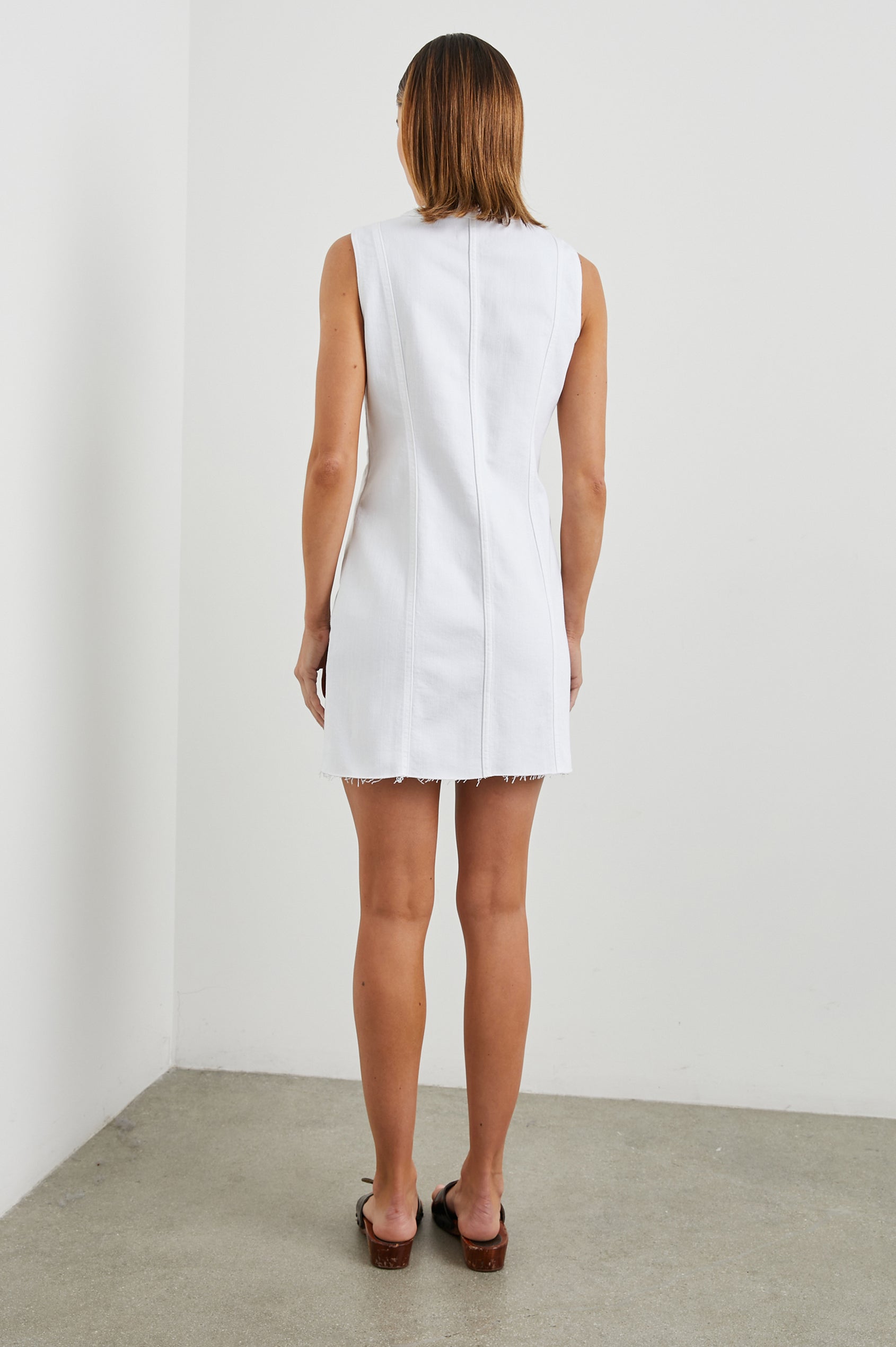 Strand Sleeveless Denim Mini Dress with a V Neck, a Raw Hem, Wide Shoulder Straps, a Row of Buttons that Extend Diagonally Across the Bodice and then Vertically Down the Skirt, Vertical Seams in Salt White Colorway - Back Full Body View Featured on Model  