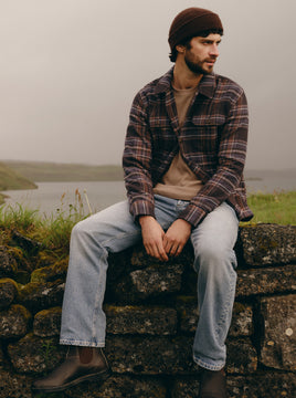 Man wearing a beige shirt under a plaid jacket with light wash jeans and boots while sitting on a stone ledge.