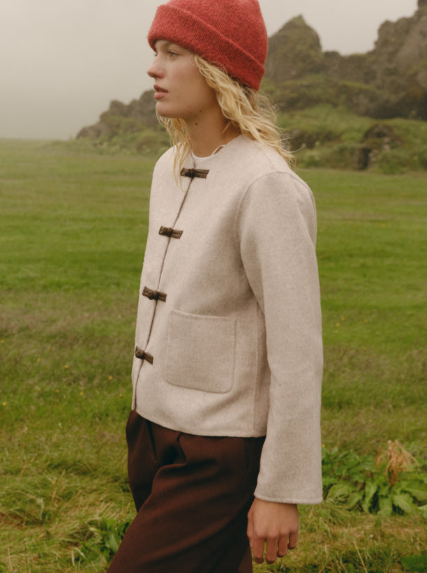 A person standing outdoors in a grassy field wearing a light beige wool-like jacket with dark toggle closures, dark brown trousers, and a red knit beanie.