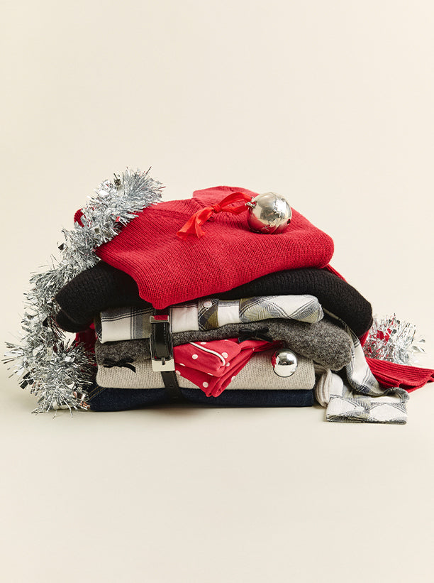 A stack of folded holiday-themed knit garments and accessories, including a bright red sweater, a black knit piece, plaid and gray patterned items, a red bandana, and silver tinsel decorations, arranged on a warm cream background.
