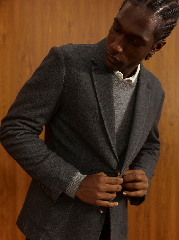 Man wearing a grey blazer with a grey sweater posed against a brown wooden background