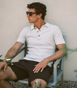 Man wearing sunglasses and a light-colored polo shirt sitting on a chair against a plain wall.