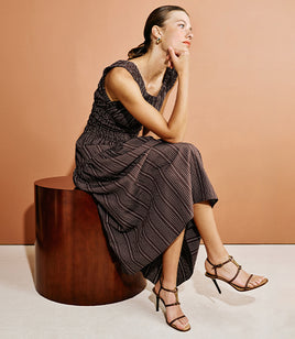 A woman sits on a dark wooden stool against a warm peach studio backdrop, wearing a sleeveless dark midi dress with thin vertical pinstripes and strappy high-heeled sandals.