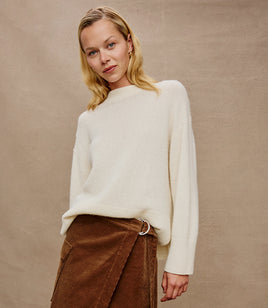 Woman wearing a white sweater and brown pants against a beige background