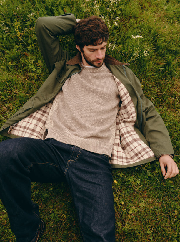 A man lying on green grass wearing an olive green jacket over a beige knit sweater and a brown-and-white plaid shirt, paired with dark blue jeans.