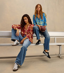 Two women sitting on metal bleachers in a studio setting, wearing plaid flannel shirts and blue jeans with sneakers. One woman holds a football.