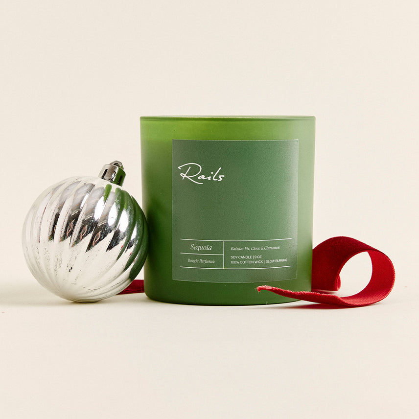 Green frosted "Sequoia" Rails soy candle beside a silver fluted ornament, with a red velvet ribbon curling in front.