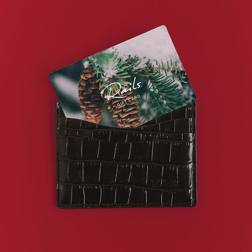 Rails gift card with a snowy pinecone image, tucked into a dark brown croc-embossed leather cardholder on a red background.