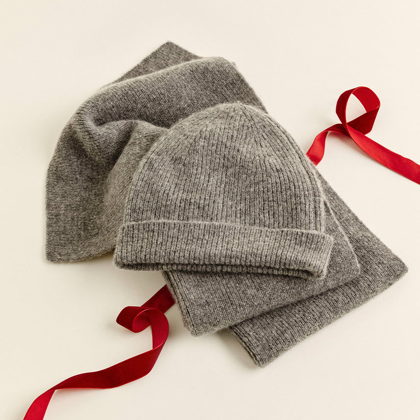 Gray ribbed knit beanie and matching scarf laid out with red satin ribbon on a cream background.