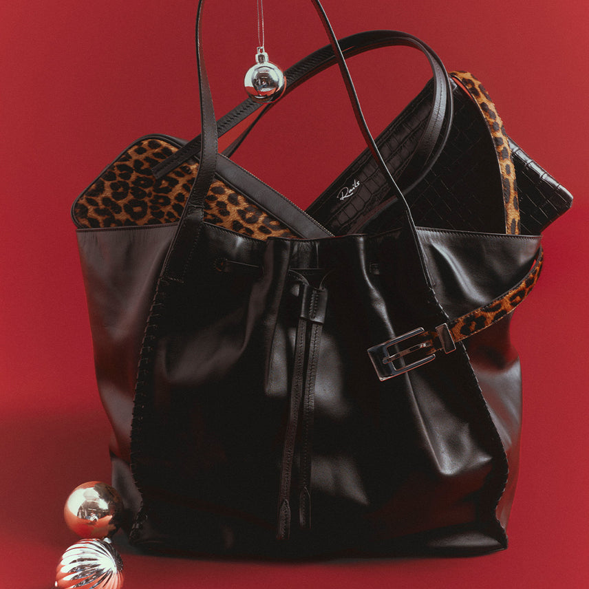 Black leather drawstring tote with whipstitched edges, holding a leopard-print calf hair tote and a black woven leather clutch, with a leopard belt draped over the front and silver ornaments against a red backdrop.