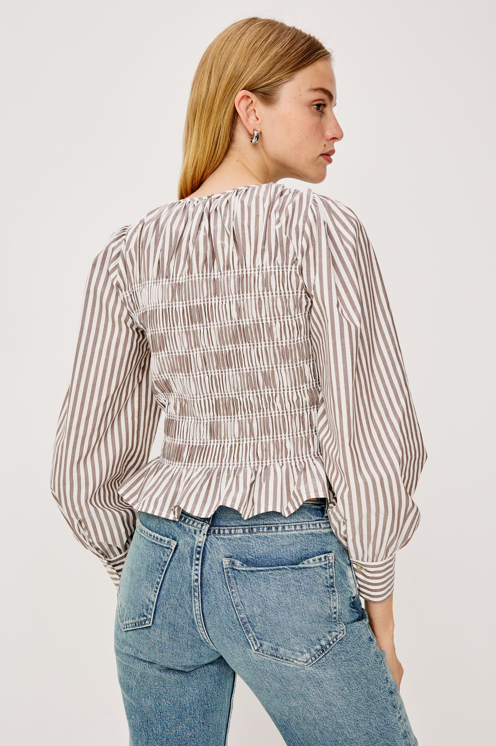 Tabi is a long sleeve crew neck top with puff sleeves and buttons at cuff, the bodice is pleated with a peplum hem in an Espresso Ivory Stripe colorway. Back view featured on model.